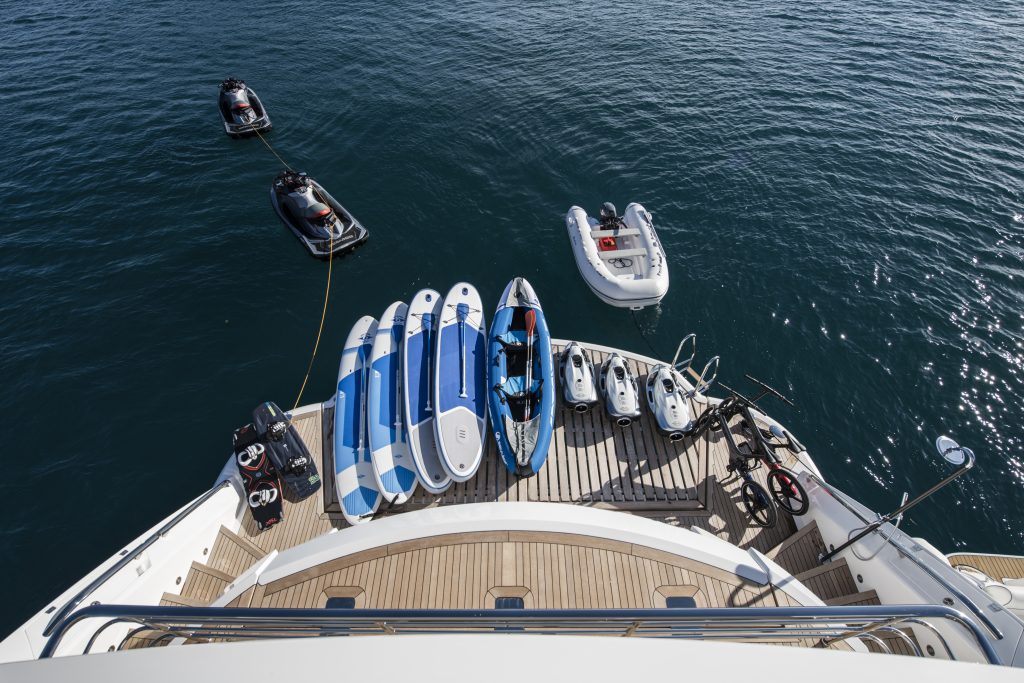 A yacht’s stern deck displays several paddleboards, a kayak, two jet skis, and bikes, with jet skis and an inflatable dinghy floating on calm blue water nearby.