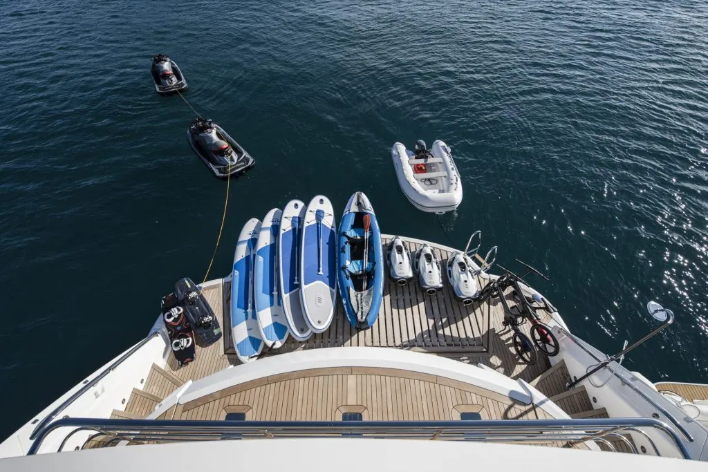 A yacht’s stern deck displays several paddleboards, a kayak, two jet skis, and bikes, with jet skis and an inflatable dinghy floating on calm blue water nearby.