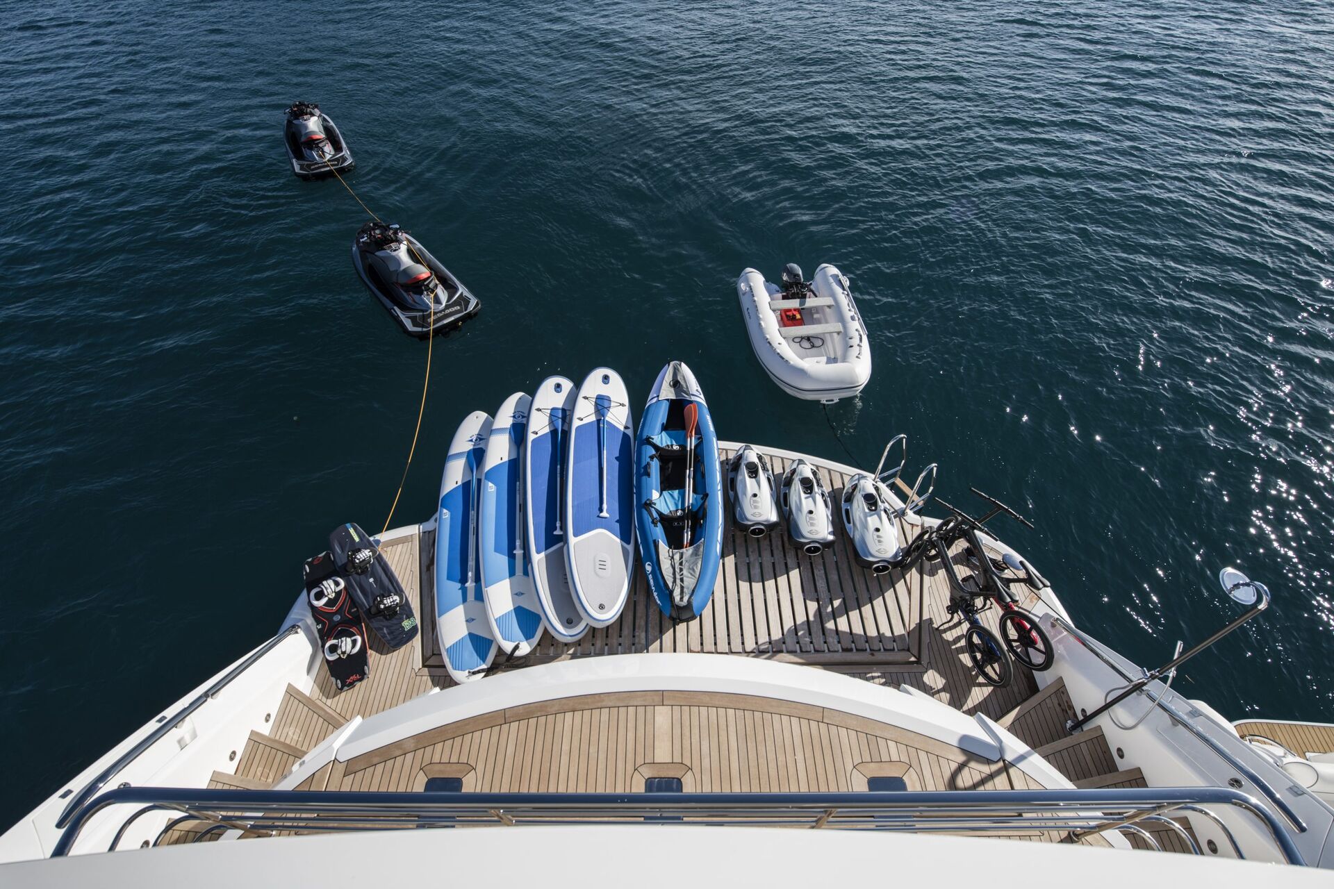 A yacht’s stern deck displays several paddleboards, a kayak, two jet skis, and bikes, with jet skis and an inflatable dinghy floating on calm blue water nearby.