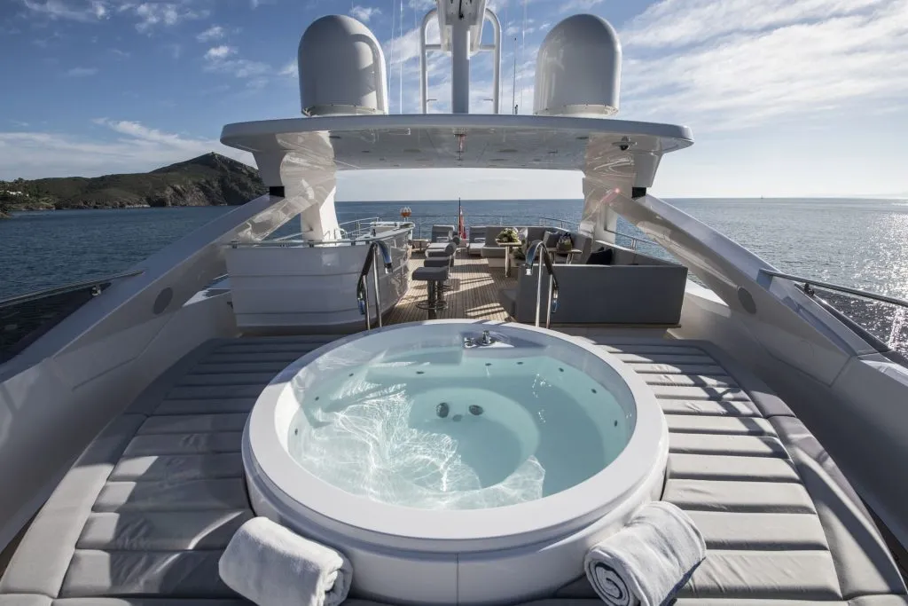 A luxurious yacht deck features a circular hot tub surrounded by sun loungers and rolled towels, with ocean views and a distant coastline under a partly cloudy sky.