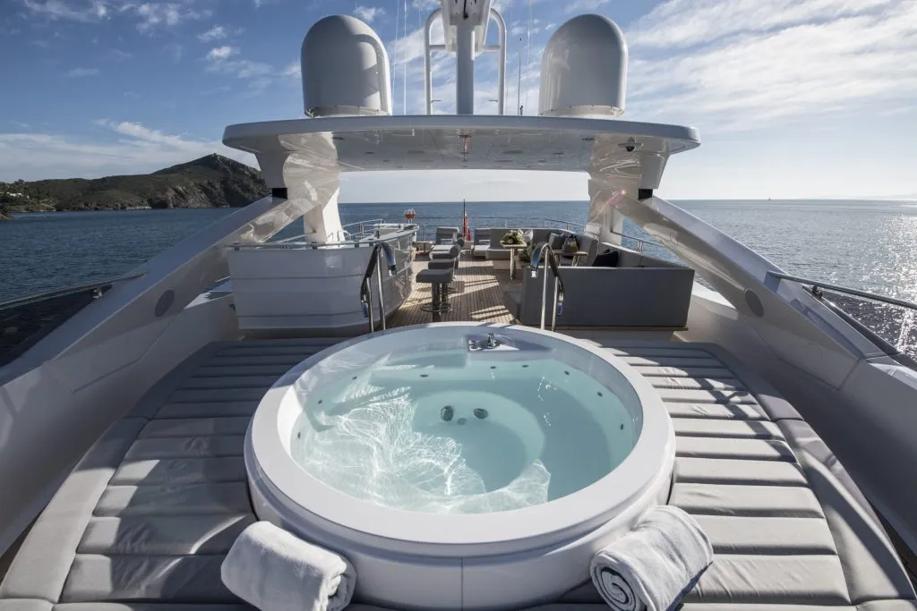 A luxurious yacht deck features a circular hot tub surrounded by sun loungers and rolled towels, with ocean views and a distant coastline under a partly cloudy sky.