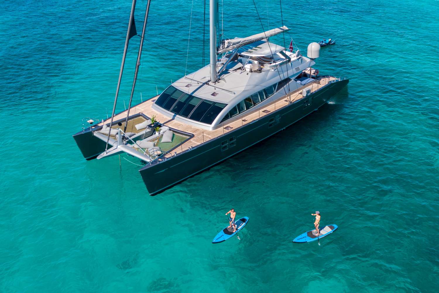 Aerial view of a luxury yacht anchored in clear turquoise water, with two people paddleboarding nearby on calm seas under bright sunlight.