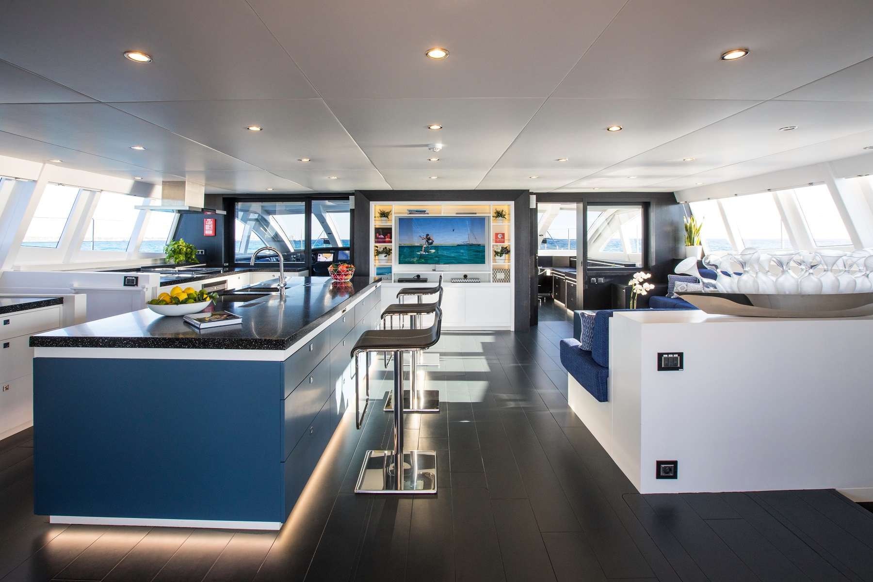 Modern yacht interior with open kitchen, island with barstools, dining area, and lounge seating. Large windows bring in natural light, and a TV is visible in the background. Surfaces are sleek and decor is minimal.