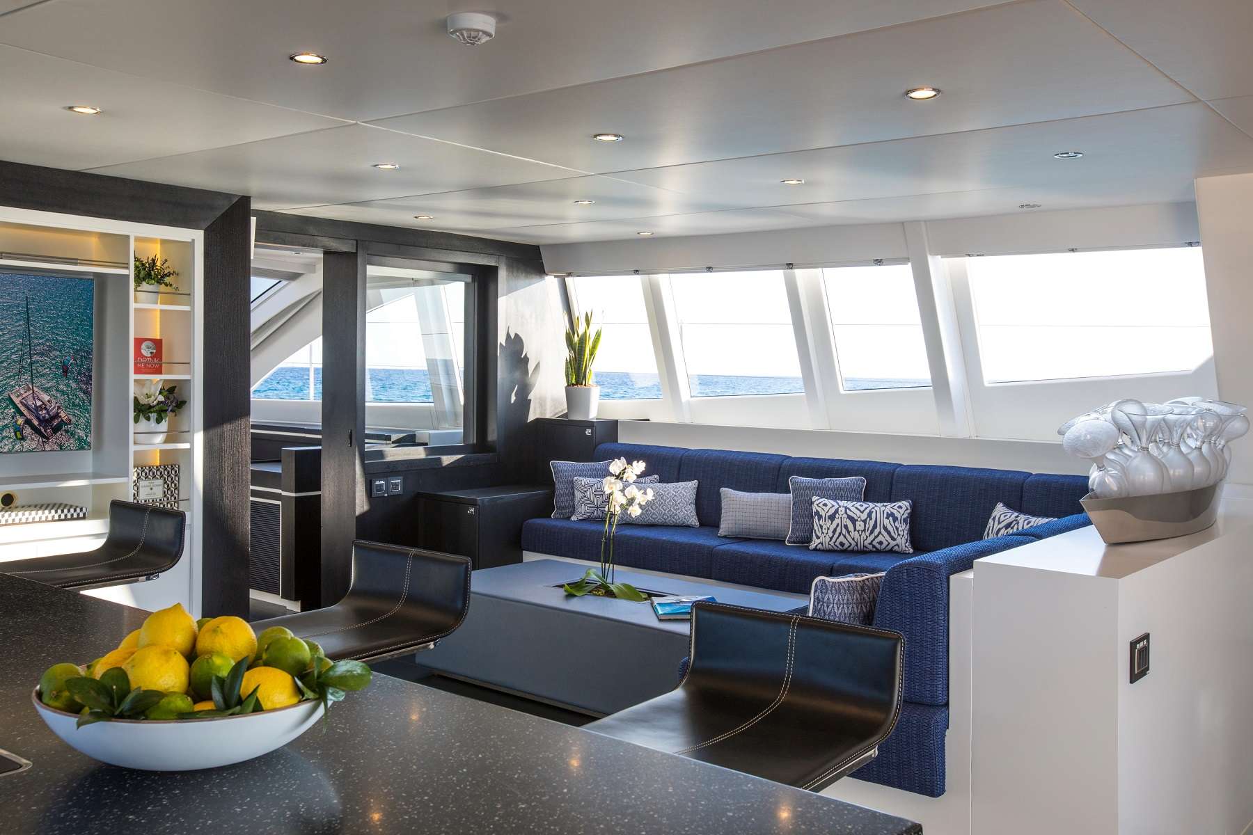 Modern yacht interior with a sunlit lounge, blue sectional sofa, decorative pillows, and large windows showing an ocean view. A bowl of lemons and limes sits on a black counter in the foreground.
