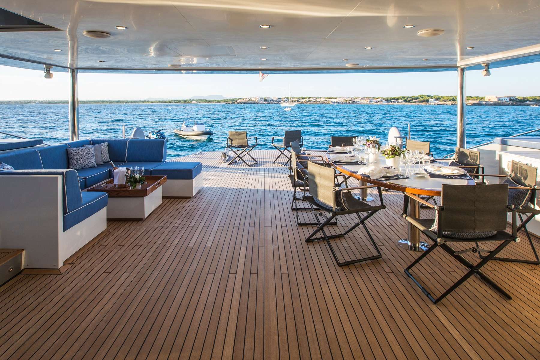 Spacious yacht deck with wooden flooring, a blue cushioned lounge area, and a dining table set for a meal. The deck overlooks calm blue water with a distant coastline and boats.