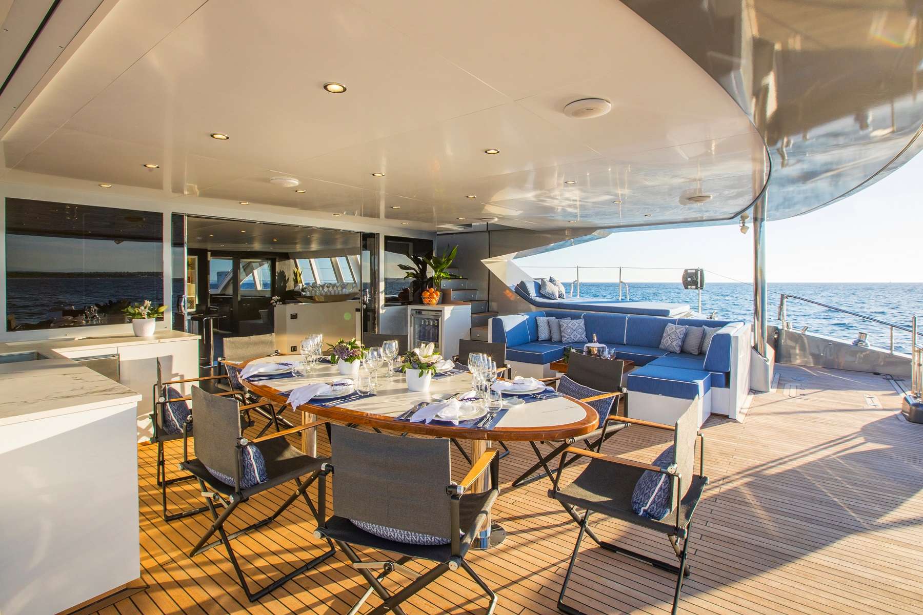 A luxurious yacht deck features a dining table set for six, cushioned seating, and elegant décor, all overlooking the ocean under a clear, sunny sky.