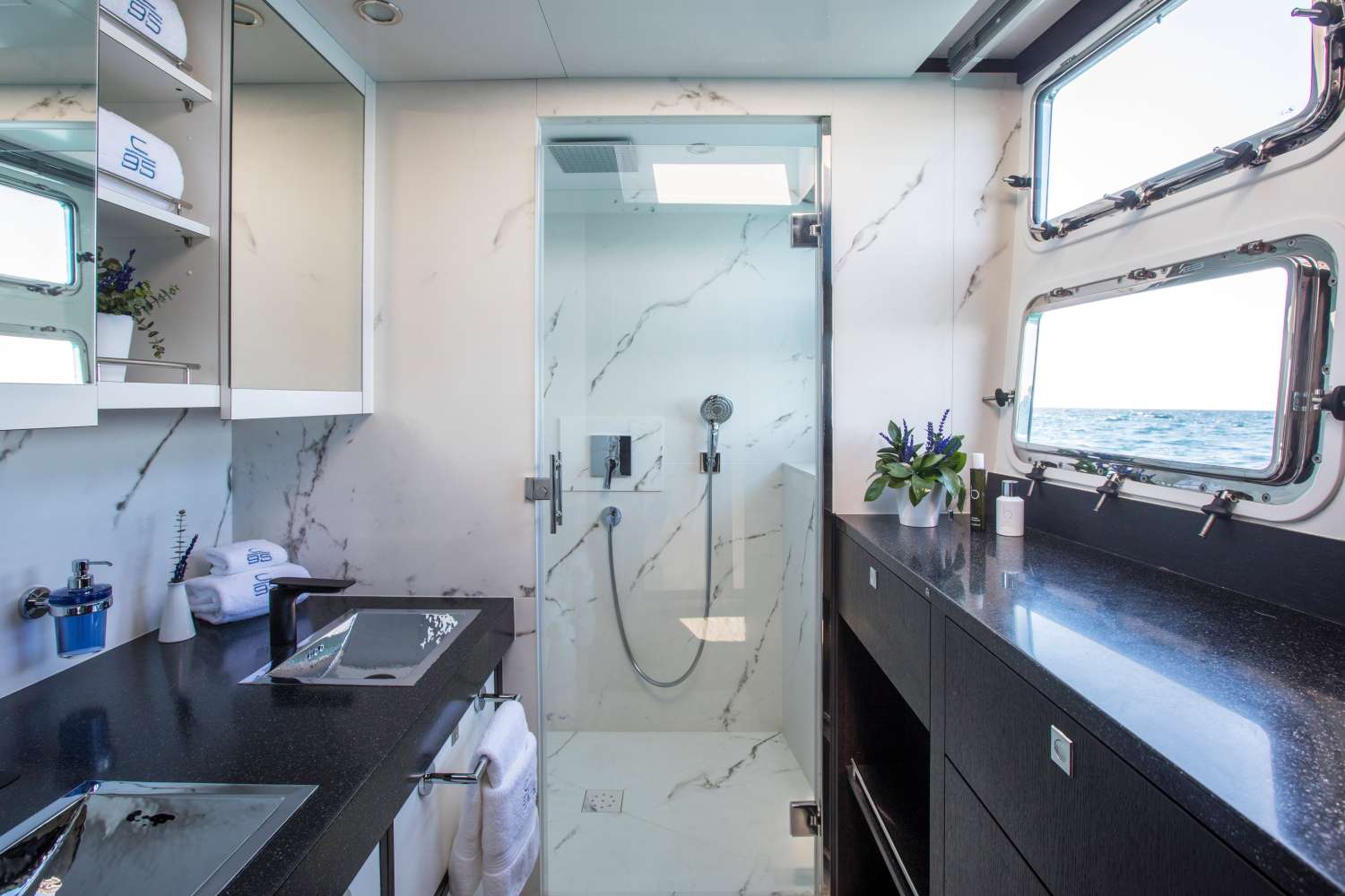 Modern yacht bathroom with white marble shower, black countertops, mirrored cabinets, and large windows showing an ocean view. The room is decorated with neatly arranged towels, toiletries, and small potted plants.