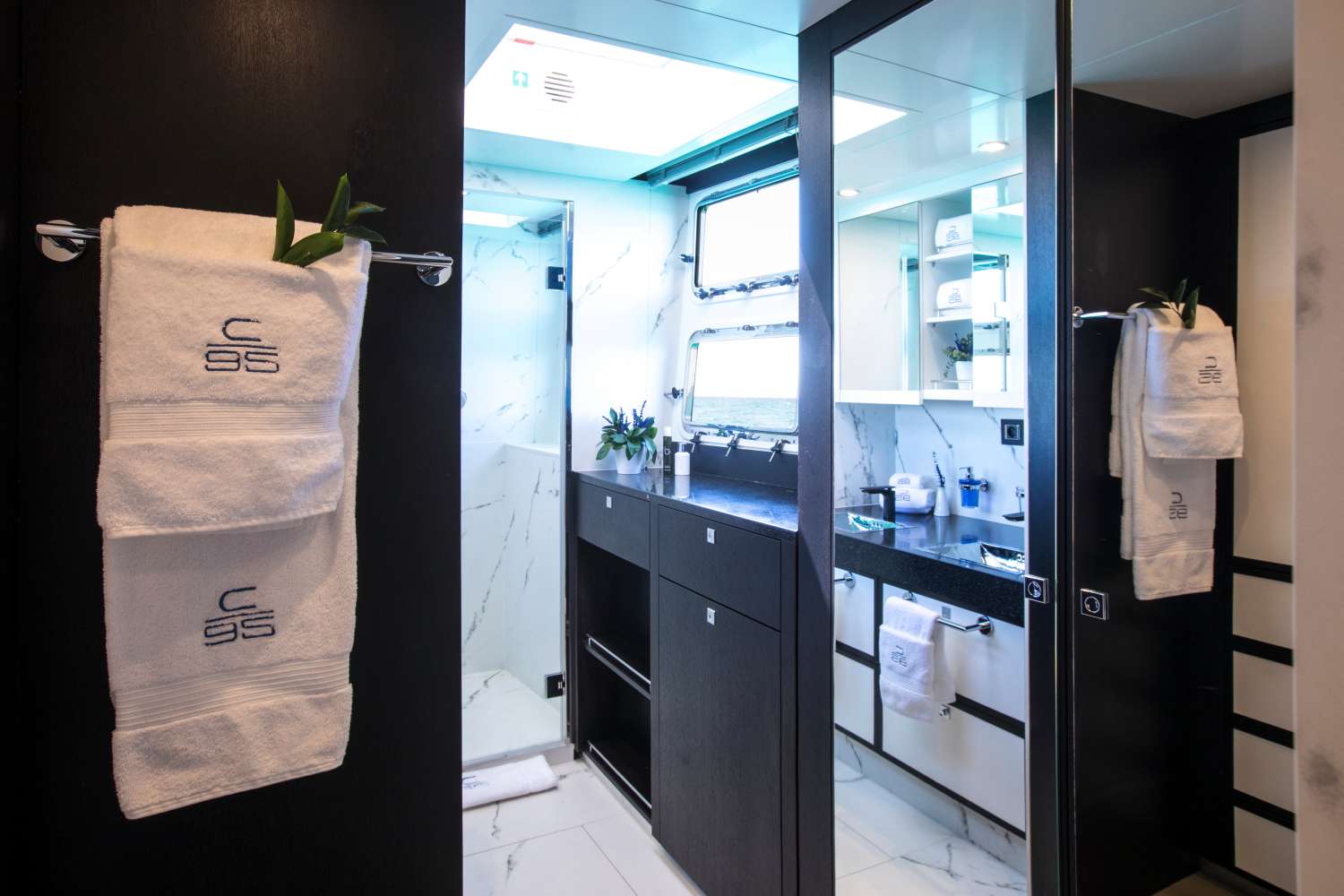 Modern yacht bathroom with white marble floor and walls, dark wood accents, double sinks, mirrors, and neatly folded white towels embroidered with C 95 hanging on the door. Small green plants decorate the space.