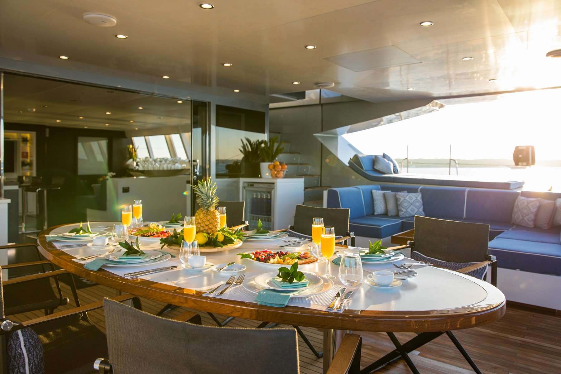 A luxurious yacht dining area set for six with plates, glasses of orange juice, fruit, and salads on a wooden table; sunlight streams in, illuminating modern seating and ocean views in the background.