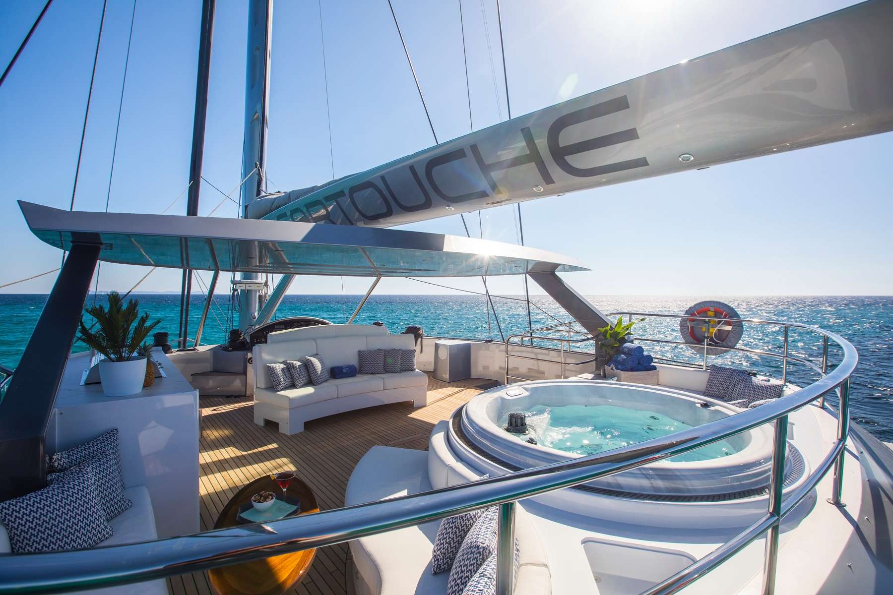 A luxurious yacht deck with a hot tub, white sofas, potted plants, and ocean views under a clear blue sky. Sunlight reflects off the water and yacht’s sails.
