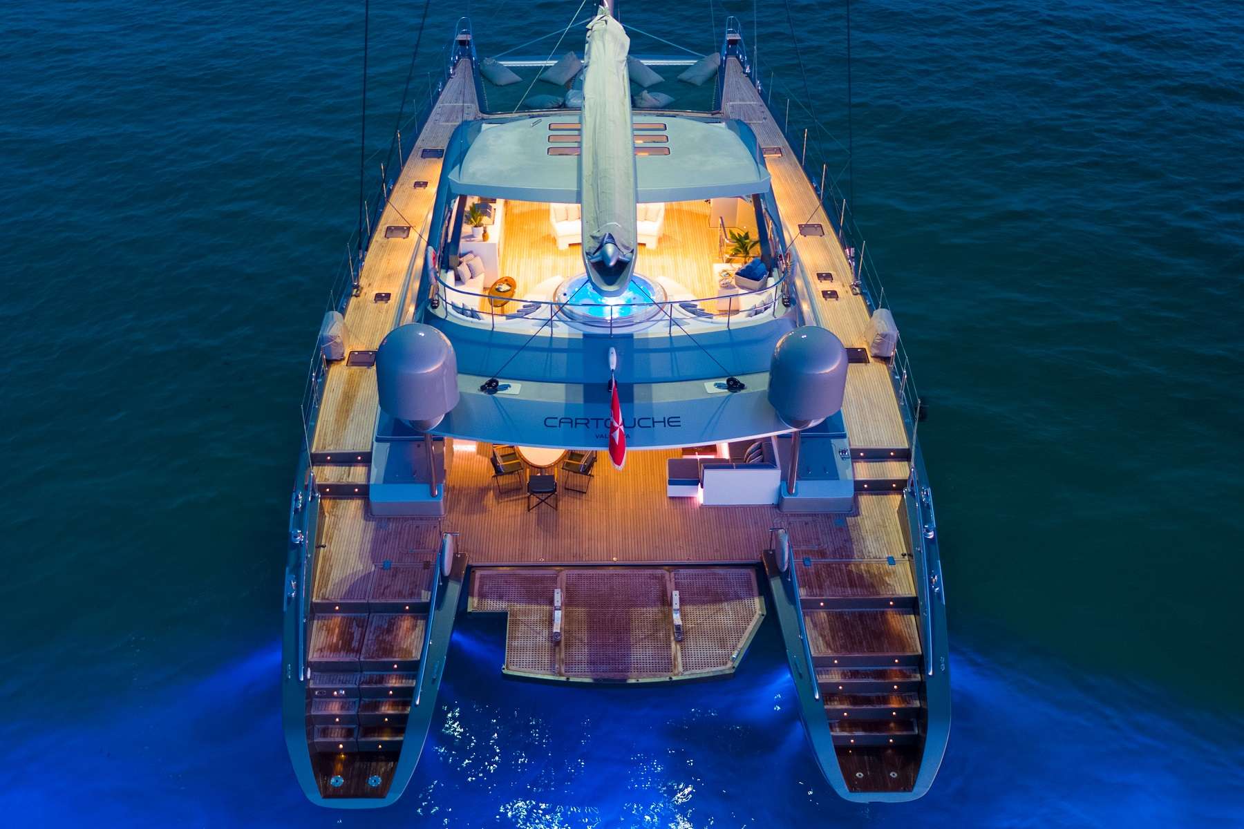 An aerial night view of a luxury catamaran yacht with deck lights glowing, illuminating the wooden decks and blue water below. The yacht, named Cartouche, is anchored and appears modern and spacious.