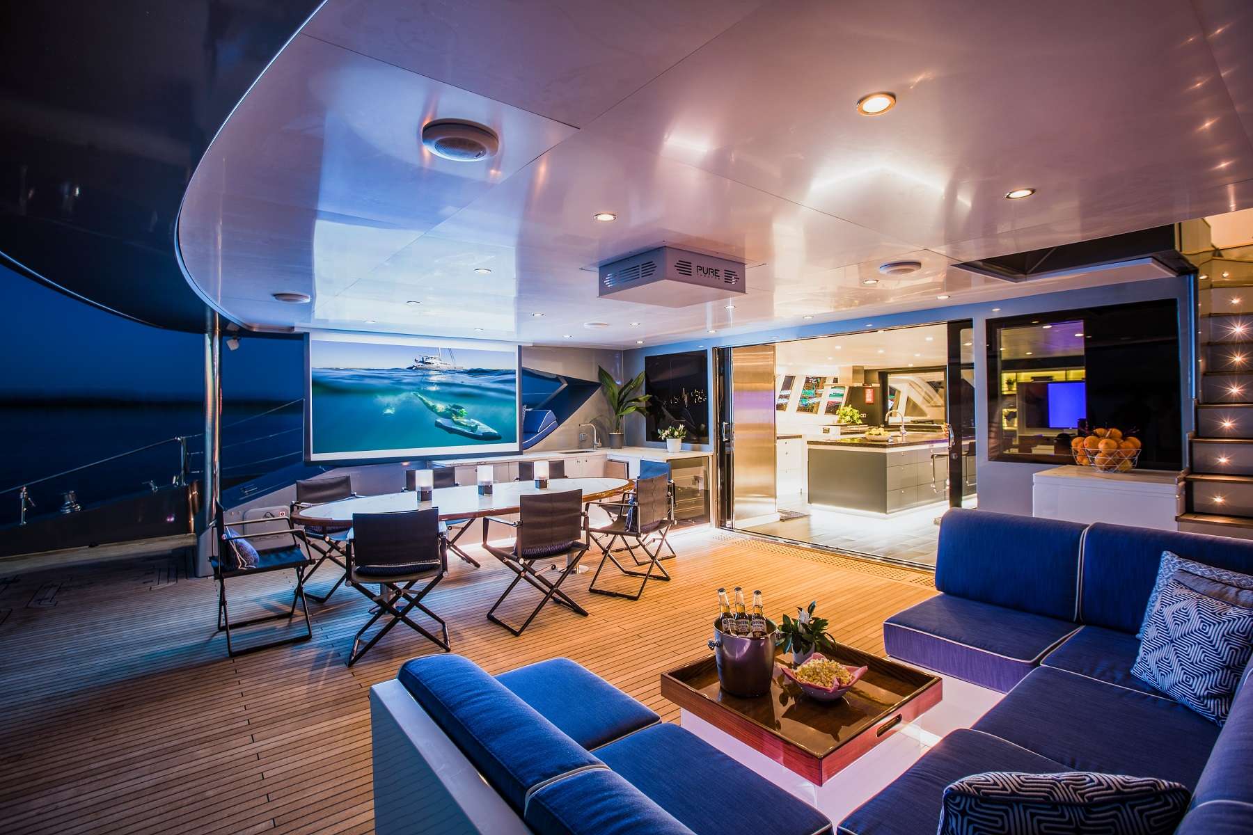 Modern yacht deck at night with a large outdoor TV, dining table and chairs, blue cushioned seating, and wooden flooring. The space is illuminated with soft lighting, creating a luxurious and inviting atmosphere.