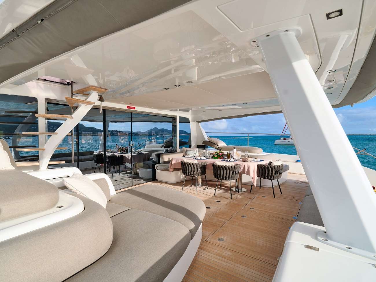 Una cubierta de yate de lujo con asientos de felpa, una mesa de comedor preparada para una comida, barandillas de cristal y una vista del mar turquesa y los barcos de fondo bajo un cielo parcialmente nublado.