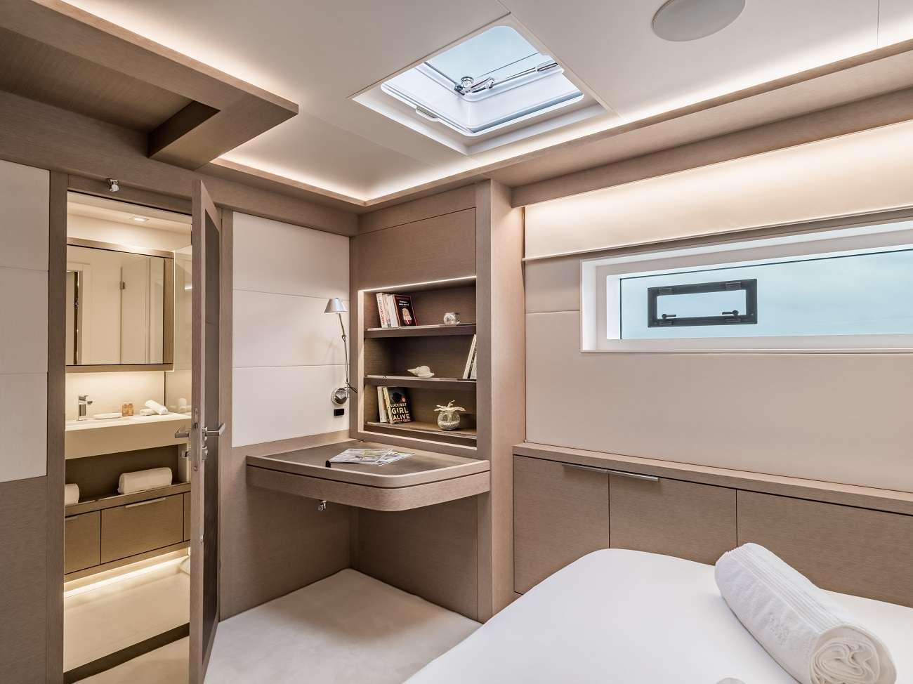 Un dormitorio moderno y minimalista con tonos beige, estanterías empotradas, un pequeño escritorio, una ventana rectangular, iluminación empotrada y un cuarto de baño adjunto visible a través de una puerta abierta. Sobre la cama hay una toalla enrollada.