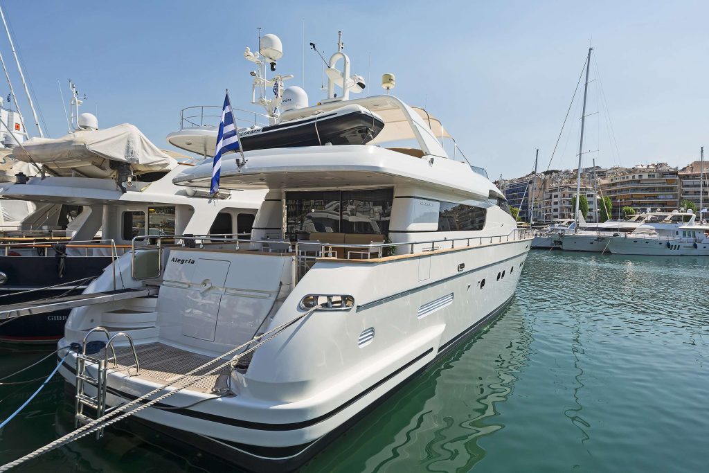 A luxury white yacht available for yacht charter is docked at a marina, flying a Greek flag. Other yachts for charter and boats are moored nearby, with buildings visible in the background under a clear blue sky.
