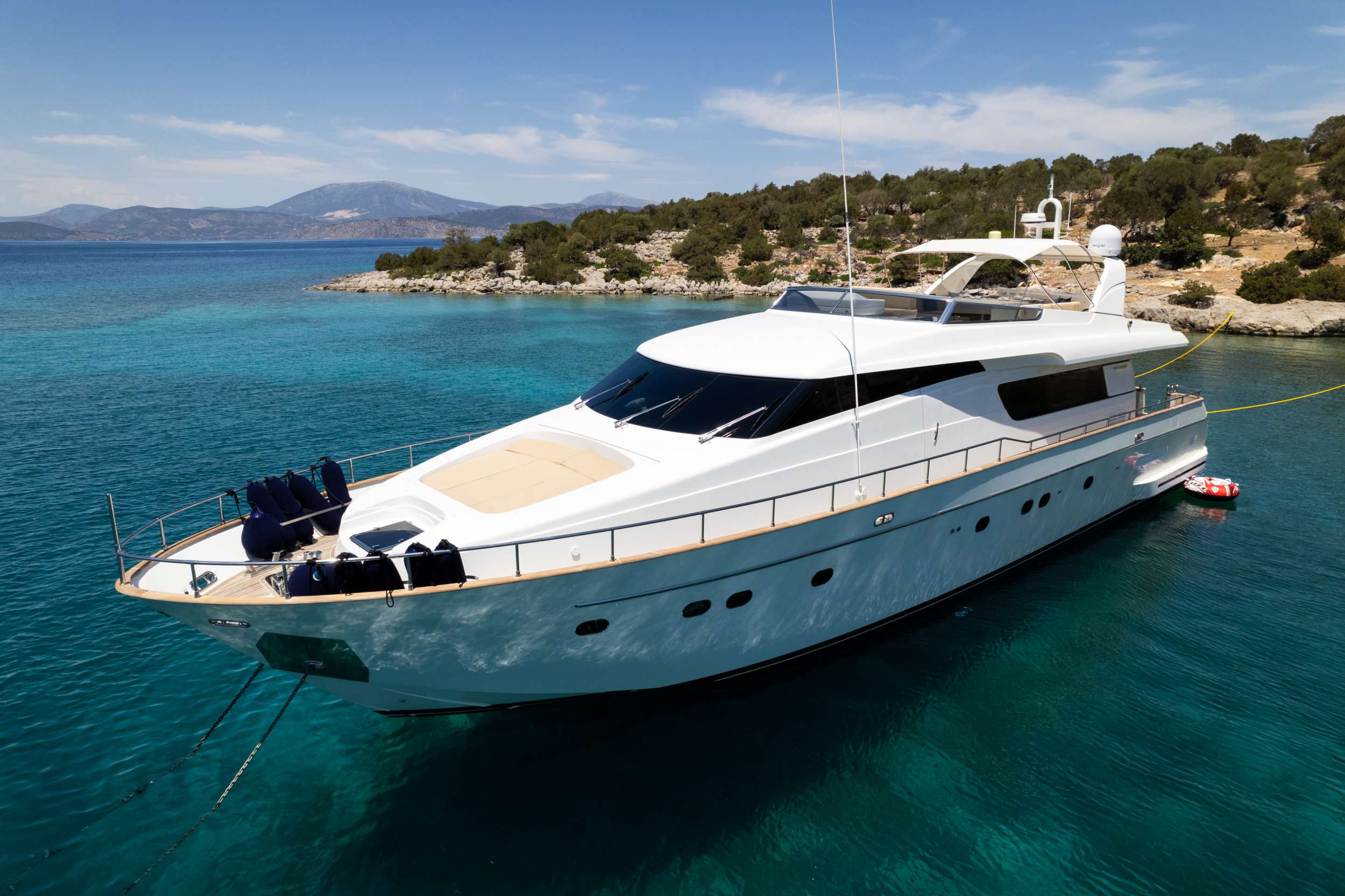 A large white luxury yacht, available for yacht charter, is anchored in clear turquoise water near a rocky, tree-lined shoreline under a sunny blue sky with distant mountains in the background.