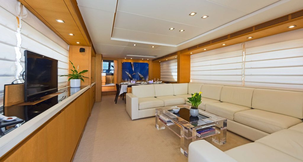 A luxurious yacht charter interior featuring cream-colored sectional sofas, a glass coffee table, TV, indoor plants, large windows with blinds, and a dining area in the background. The modern space is bright and inviting.