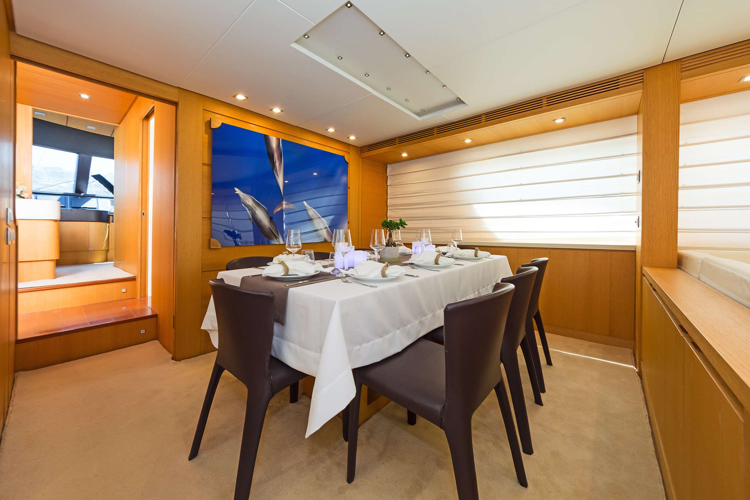A modern yacht dining room with a rectangular table set for six, brown chairs, white tablecloth, wooden walls, large window with blinds, and a vibrant blue artwork—perfect for enjoying meals during your luxury yacht charter.