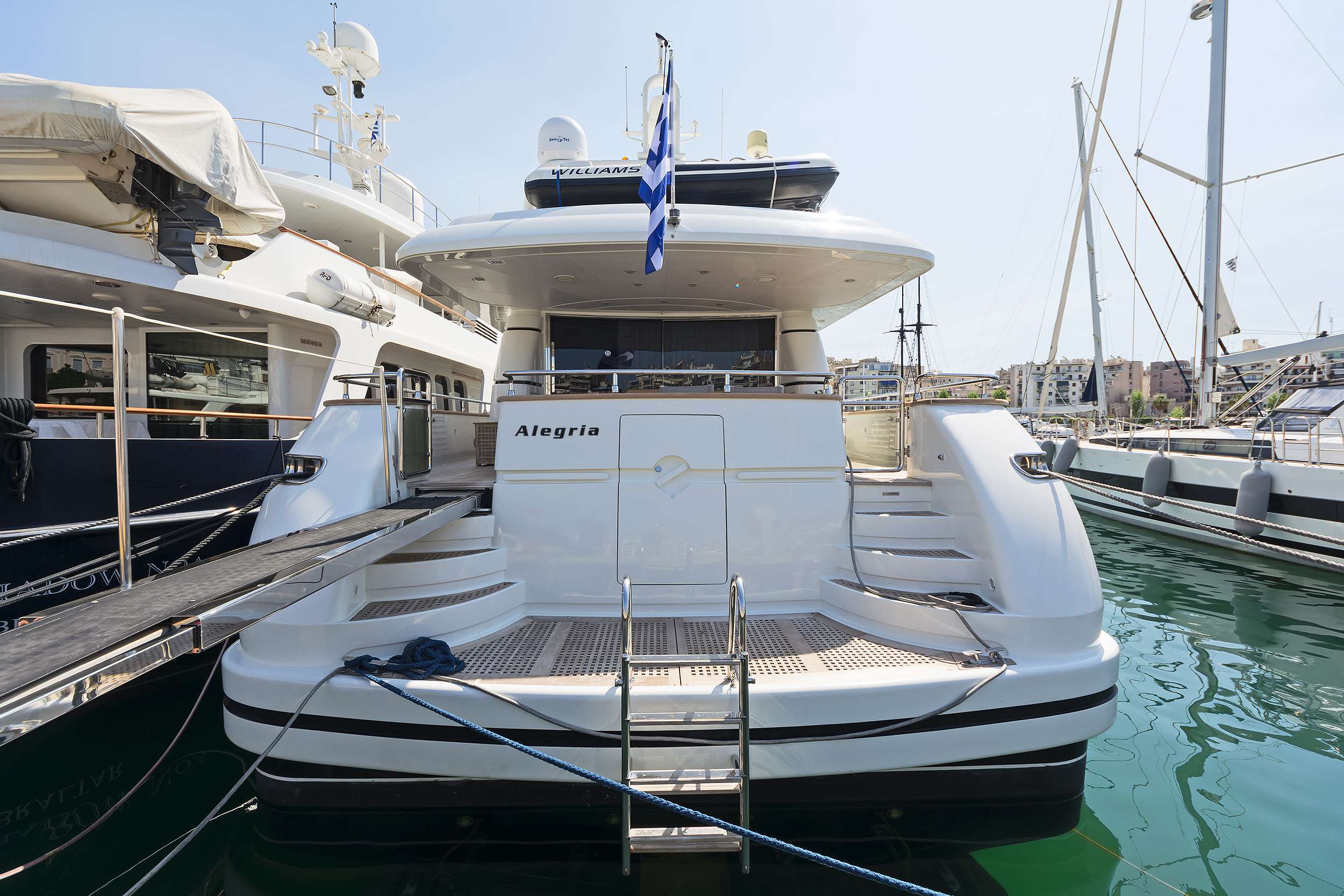 A luxury white yacht named Alegria is docked at a marina, with a metal gangway extending to the left. The calm water and nearby yachts for charter create an inviting atmosphere for your next yachting adventure.