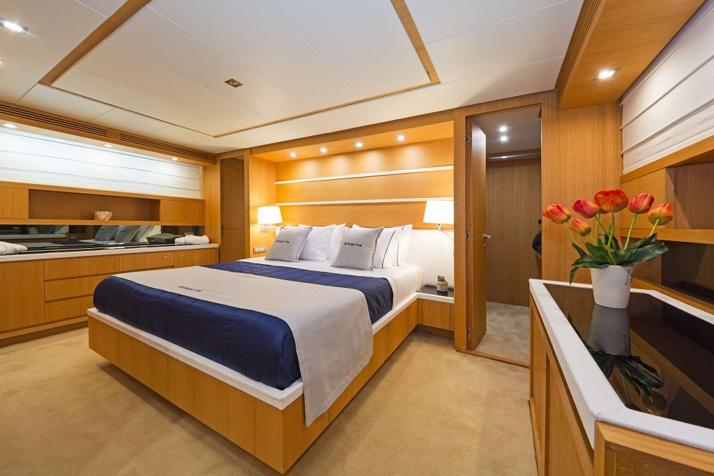 Modern yacht bedroom with wood paneling, a large bed with navy and beige bedding, built-in storage, soft lighting, a mirror, and a vase of red tulips—ideal for your next luxury yacht rental or charter experience.