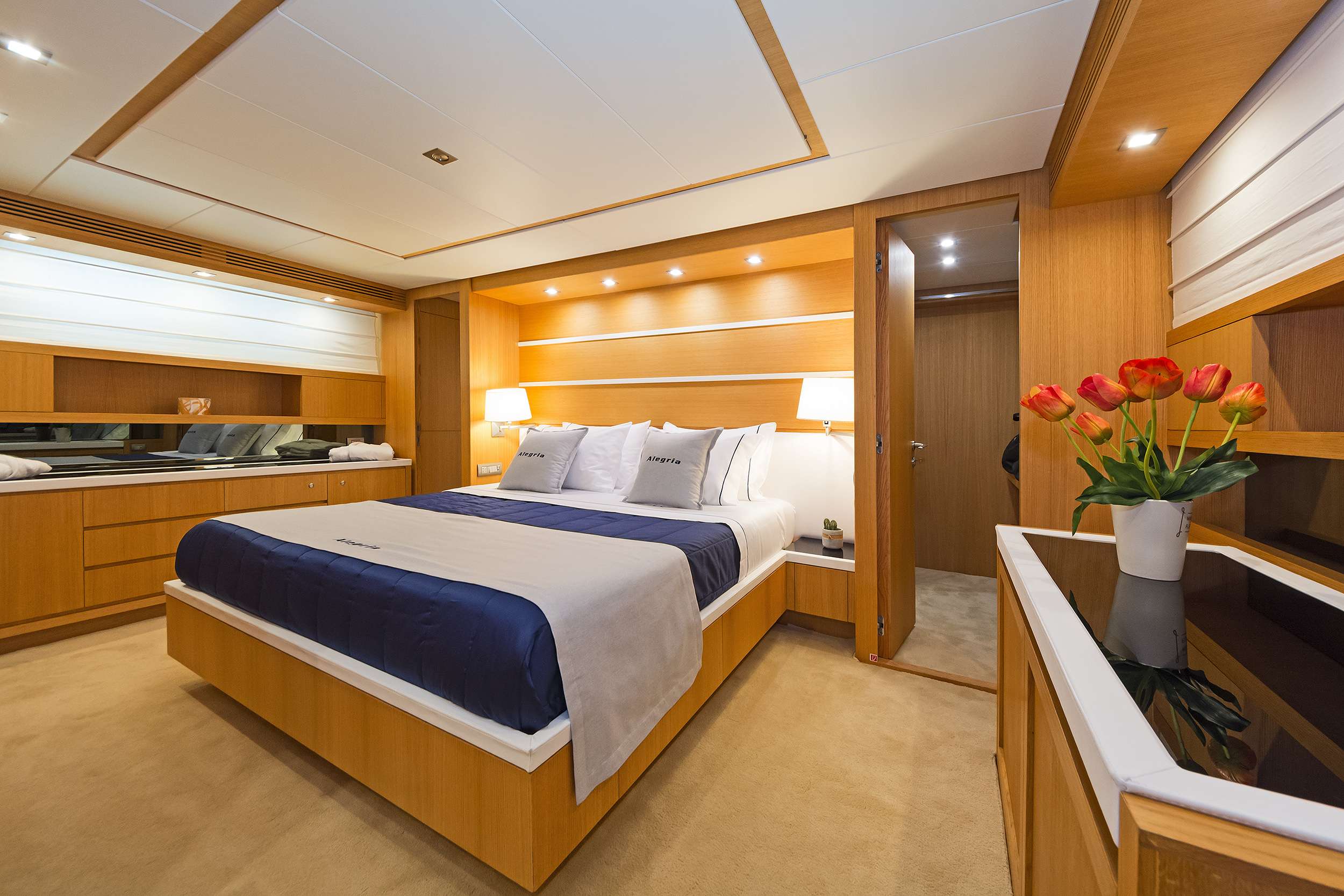 Modern yacht bedroom with wood paneling, a large bed with navy and beige bedding, built-in storage, soft lighting, a mirror, and a vase of red tulips—ideal for your next luxury yacht rental or charter experience.