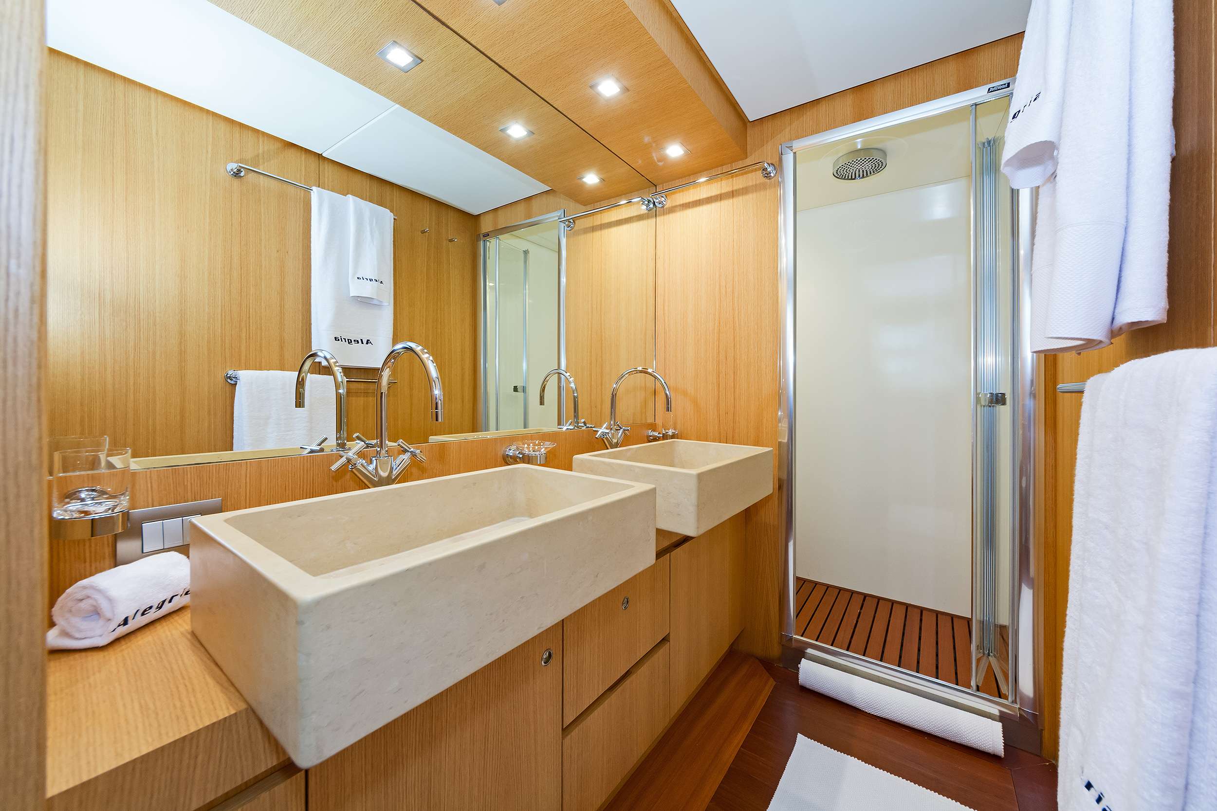 Modern bathroom with light wood walls, two beige rectangular sinks, large mirrors, and a glass shower enclosure with a rainfall showerhead—perfect luxury found on premium yacht charter experiences. White towels are neatly hung on towel racks.