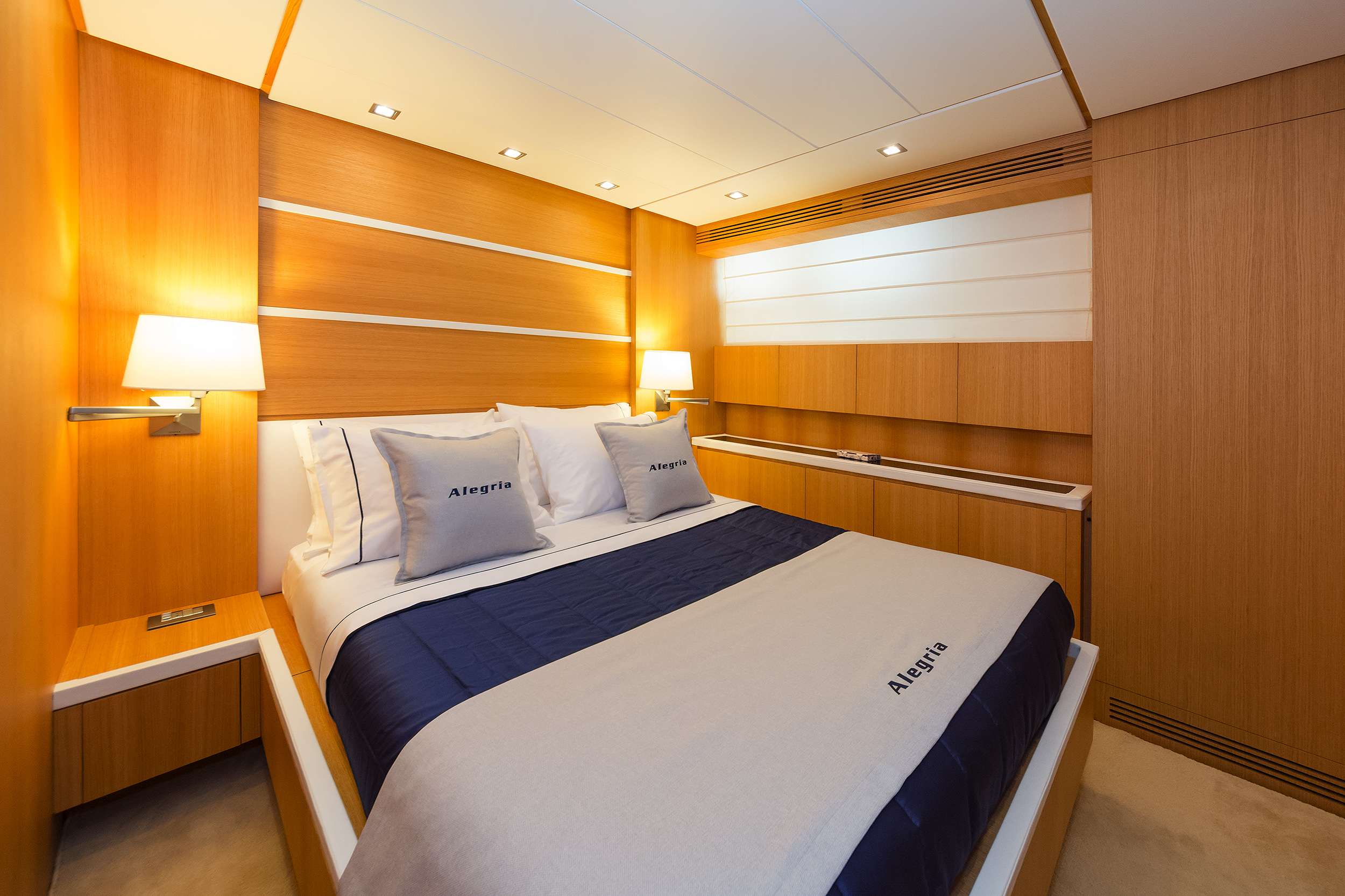 A modern yacht bedroom with wood-paneled walls, a double bed with white and navy Alegria bedding, two wall lamps, and soft overhead lighting. This clean, minimalist space is perfect for relaxing during your yacht charter experience.