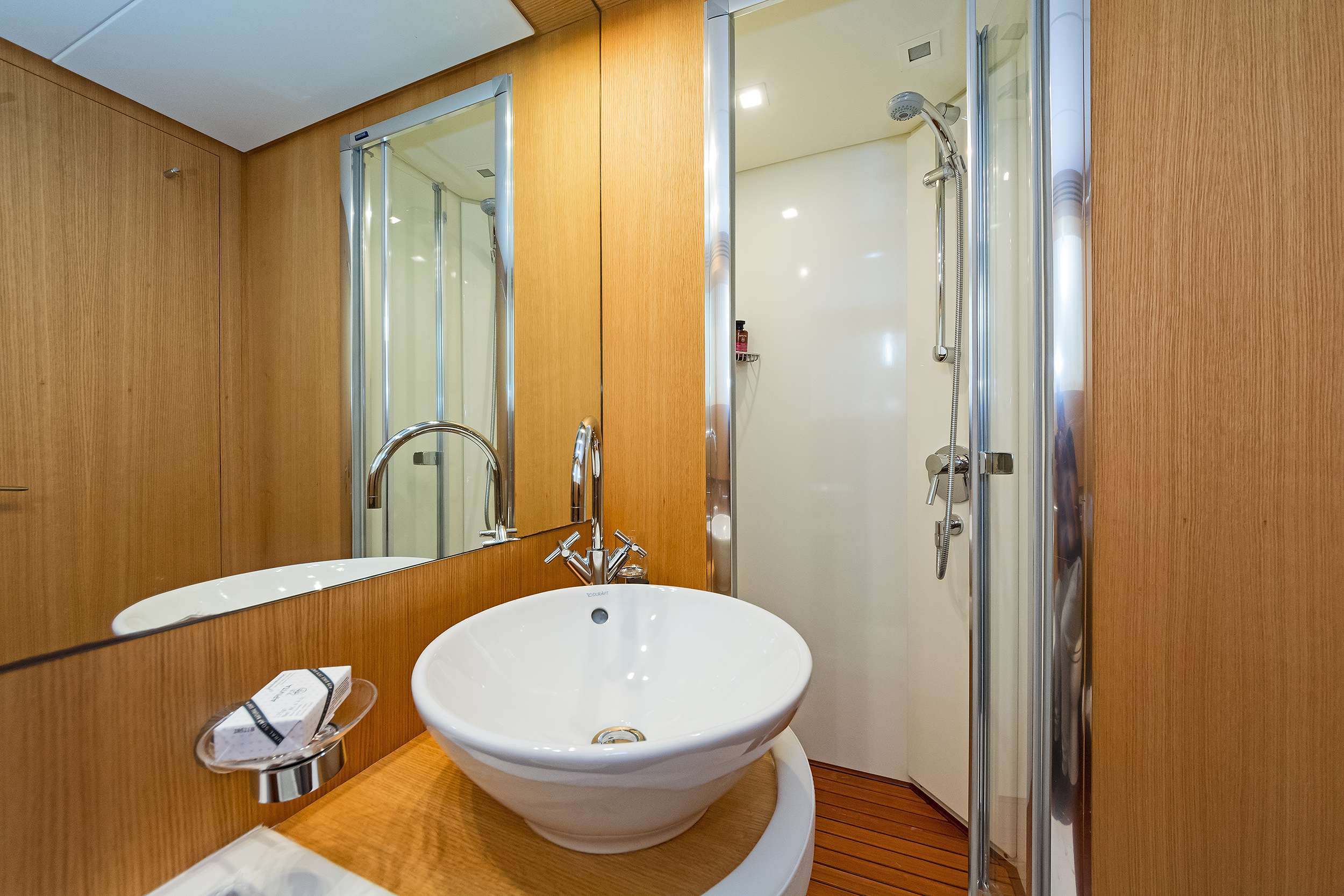 Modern bathroom with wood paneling, a round white sink on a wooden countertop, chrome faucet, wall mirror, and glass shower enclosure with chrome fixtures—perfect elegance for any luxury yacht charter experience.