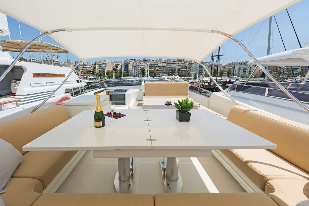 A luxurious yacht deck with a white table set for drinks and snacks, surrounded by beige cushioned seating. Perfect for a relaxing yacht charter, the scene features champagne, glasses, and marina views with other yachts for charter in the background.