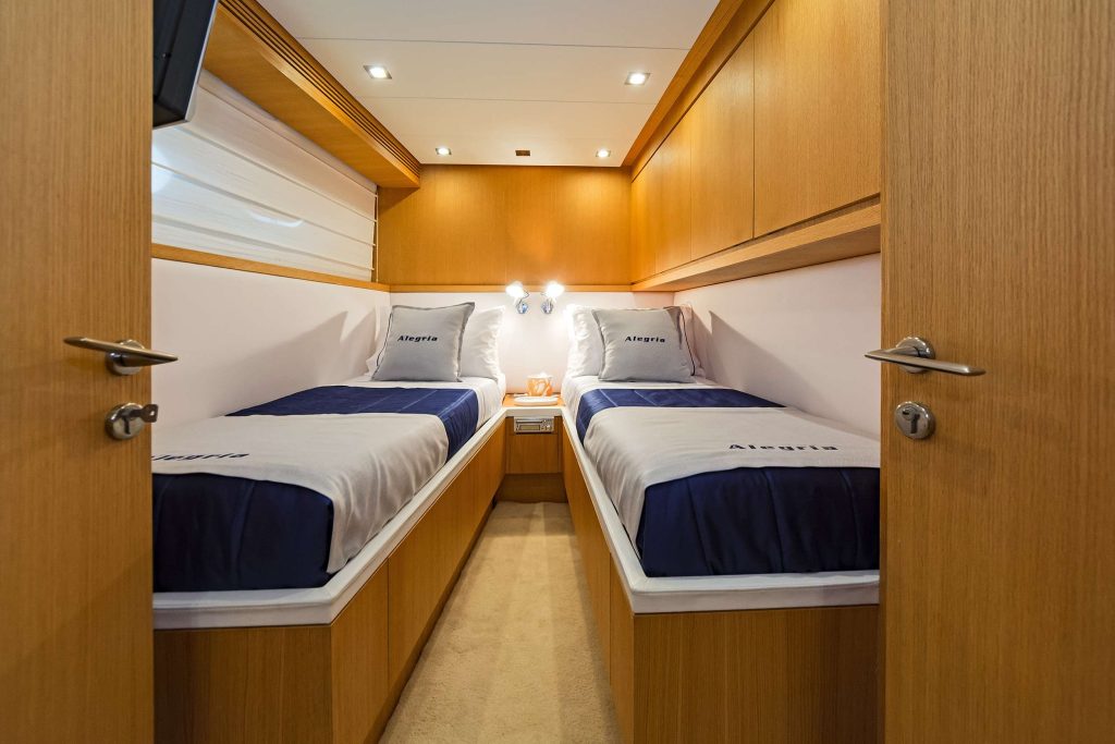 A cozy yacht cabin with two single beds, white and navy bedding, wood-paneled walls, overhead lights, and a small nightstand. Perfect for your next yacht charter, the pillows are labeled Alegria.