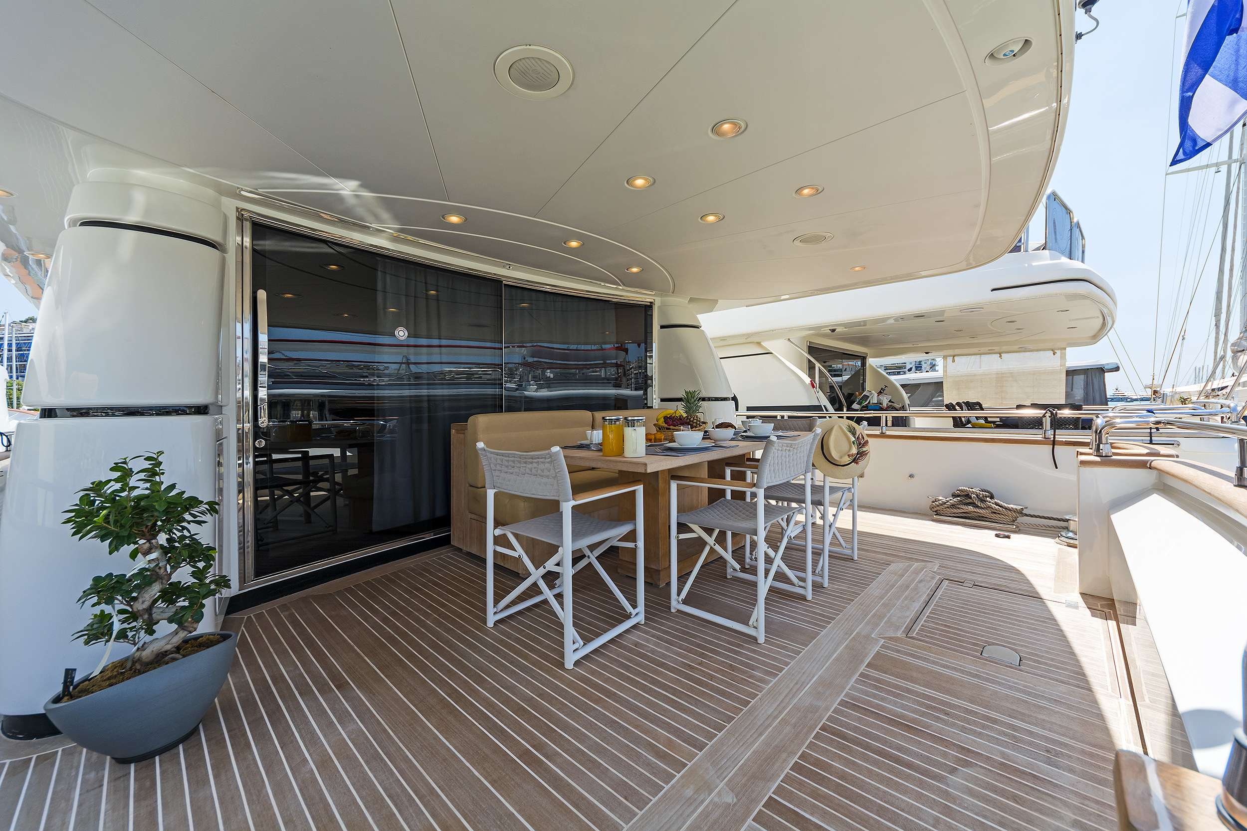 A luxurious yacht deck, perfect for yacht charter, features a wooden dining table set for four with white chairs, surrounded by potted plants under a covered ceiling with recessed lights. A marina with docked boats is visible in the background.