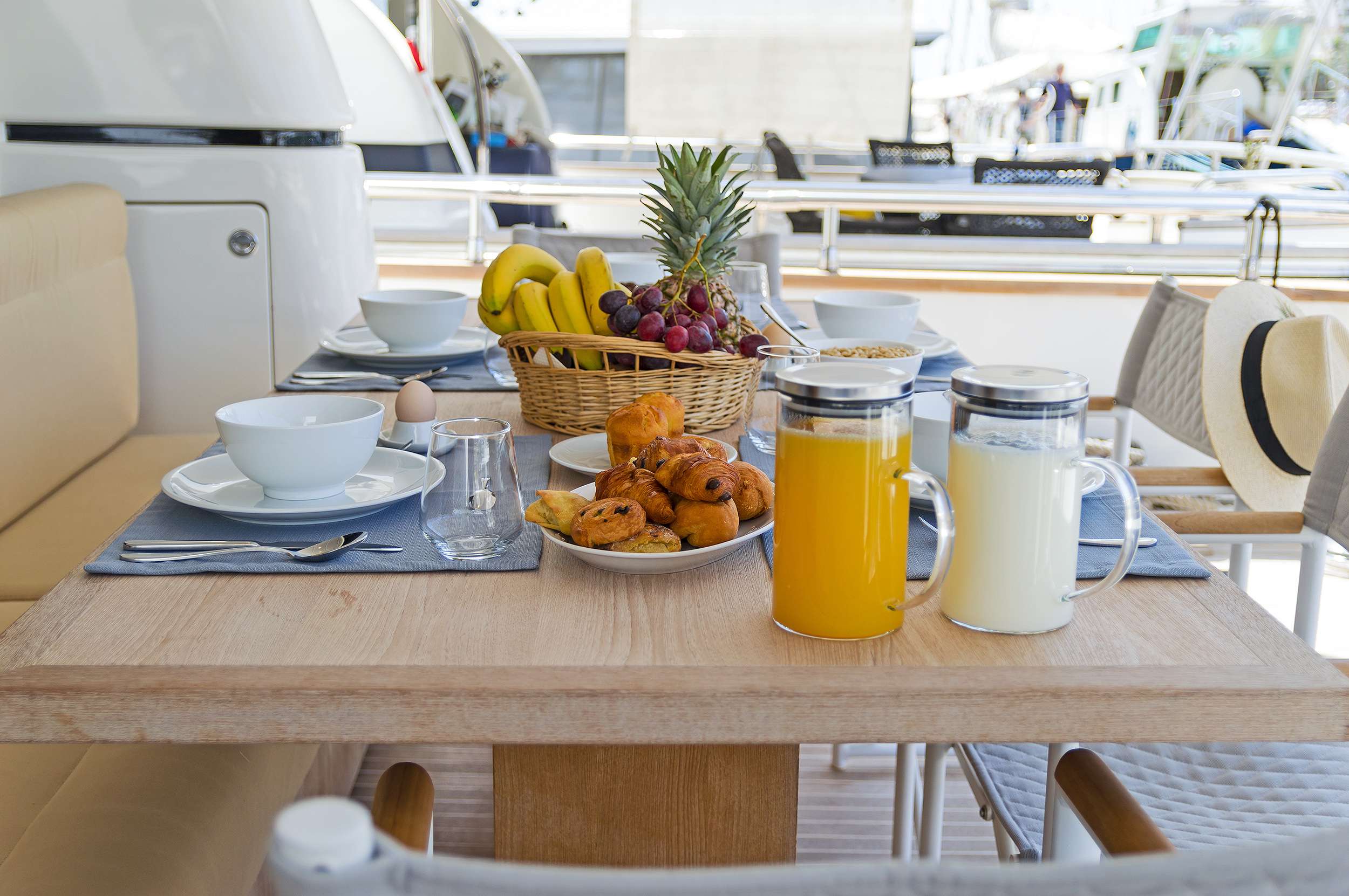 A breakfast table set on a yacht charter with bowls, pastries, fruit, glasses, a pitcher of orange juice, milk, and a sun hat resting on a chair.