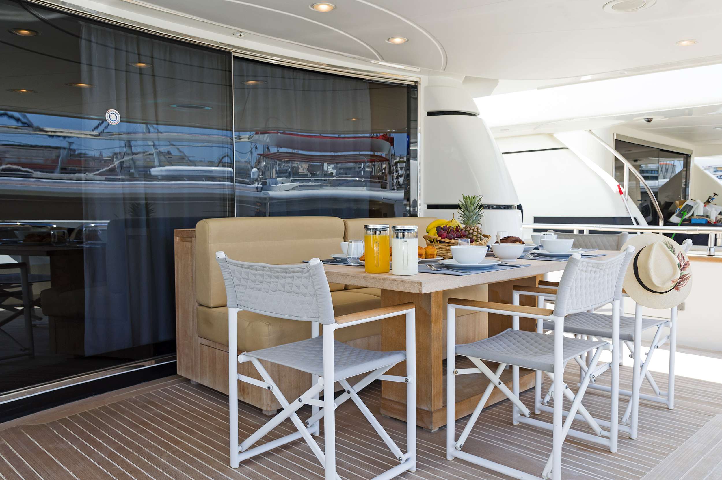 A yacht deck dining area with a wooden table set for breakfast, including juice, fruit, and coffee. Four white chairs surround the table—a perfect spot to enjoy the morning during your luxury yacht charter. Reflections of boats shimmer in the glass door behind.