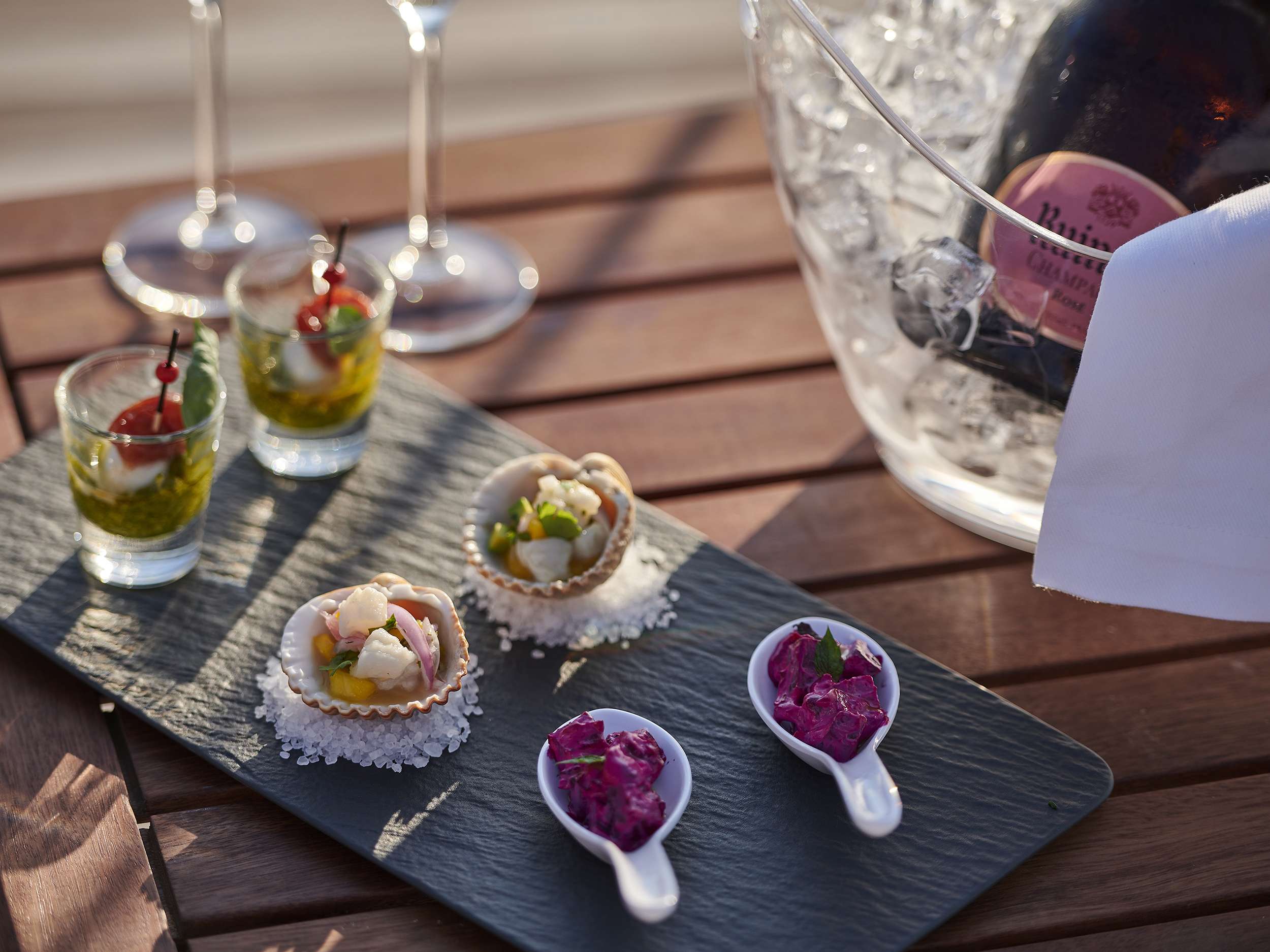 A slate tray with assorted appetizers in small shells and white spoons sits next to shot glasses with green garnish. Behind, a bucket of rosé champagne on ice hints at the luxury of yachts for charter, all set on a wooden table.