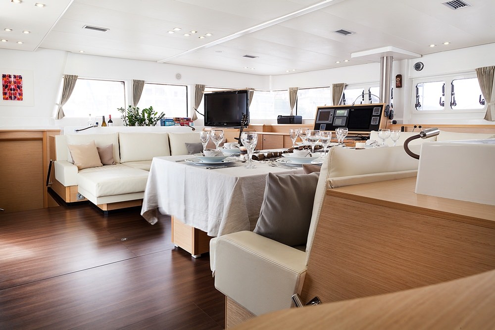 Modern yacht interior with wood flooring, cream-colored sofas, a dining table set with glassware and plates, large windows with beige curtains, and natural light filling the spacious, elegant room—perfect for a memorable yacht charter experience.