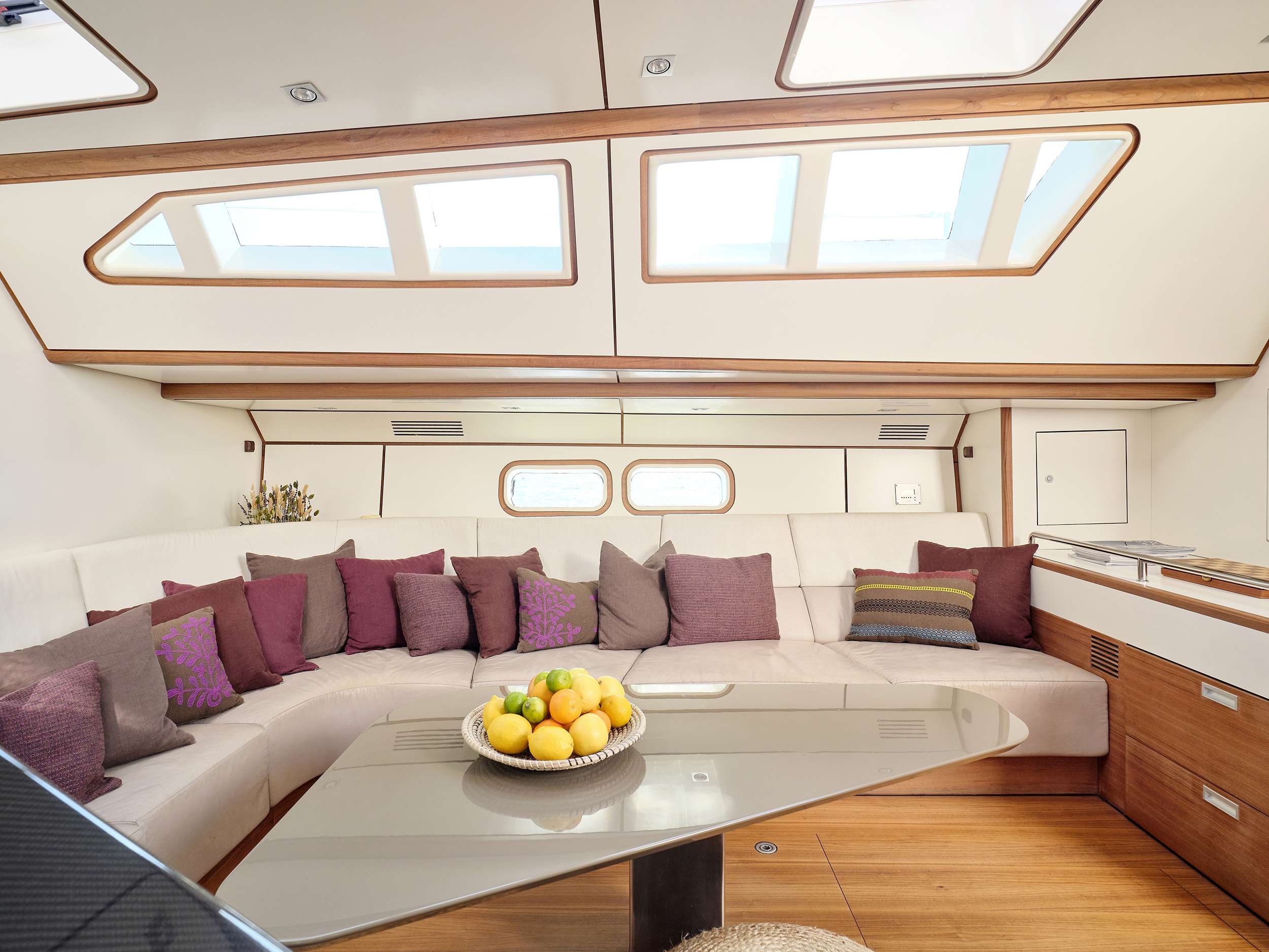 A modern yacht interior designed for yachts for charter, featuring a curved white sofa with purple cushions, a glass table with lemons, large windows, and wood accents that create a bright and inviting atmosphere.