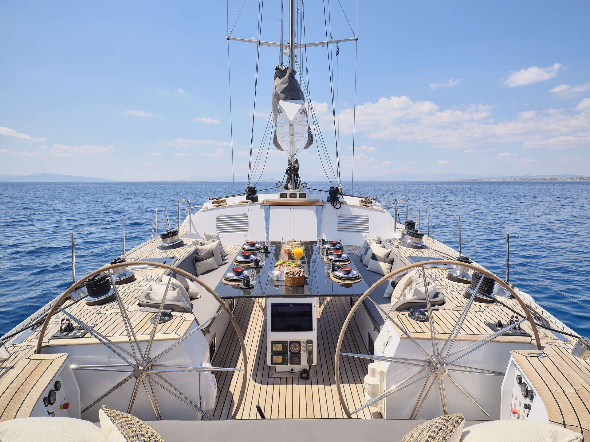A luxury sailboat deck available for yacht charter, featuring two steering wheels, an elegant dining table set for a meal, and cushioned seating—all overlooking the calm blue sea and a clear sky.