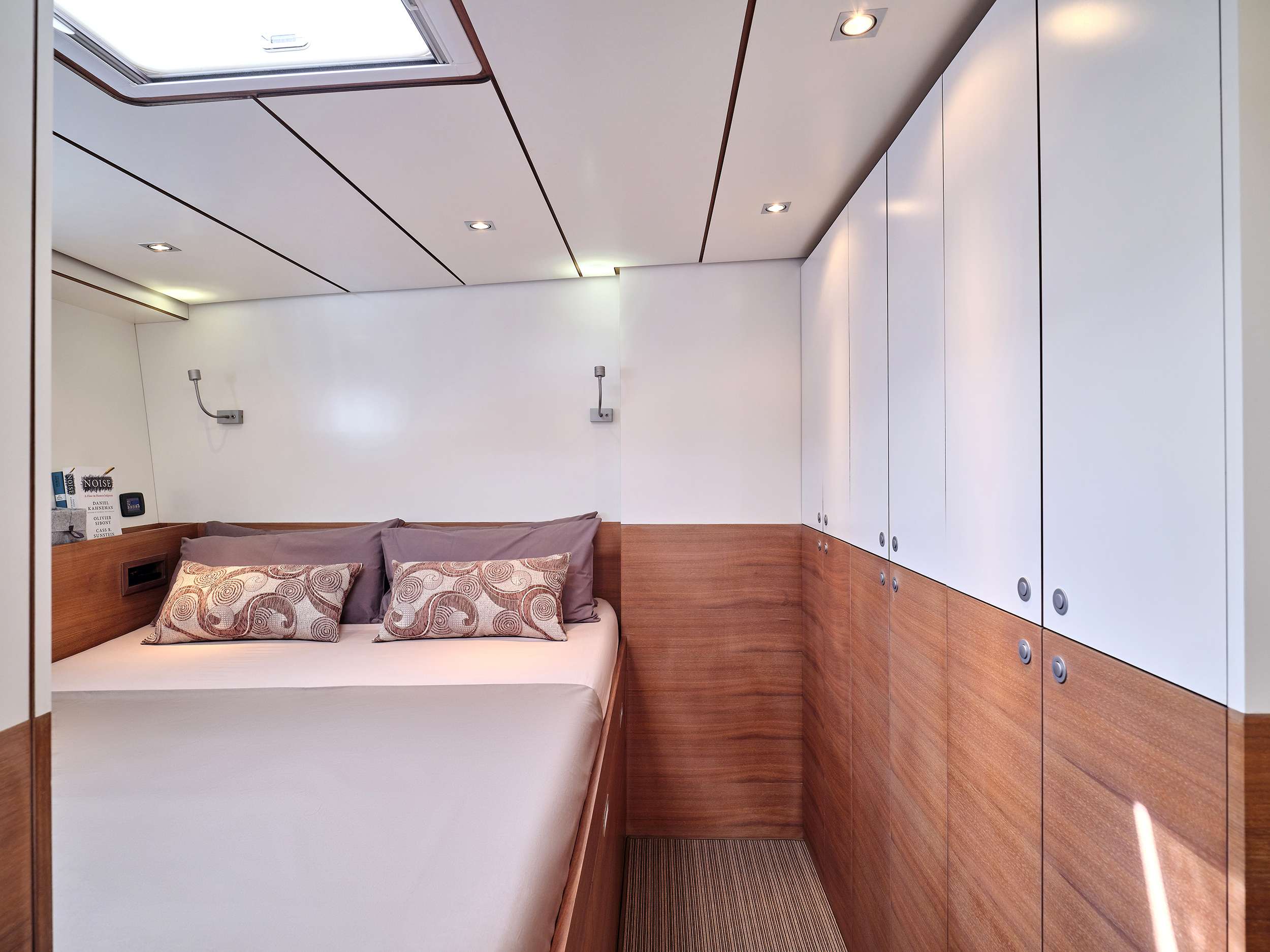 A modern yacht bedroom with a double bed, neutral bedding, decorative pillows, wood paneling, white cabinets, overhead lights, and a skylight—this inviting space is perfect for relaxing during your luxury yacht charter.