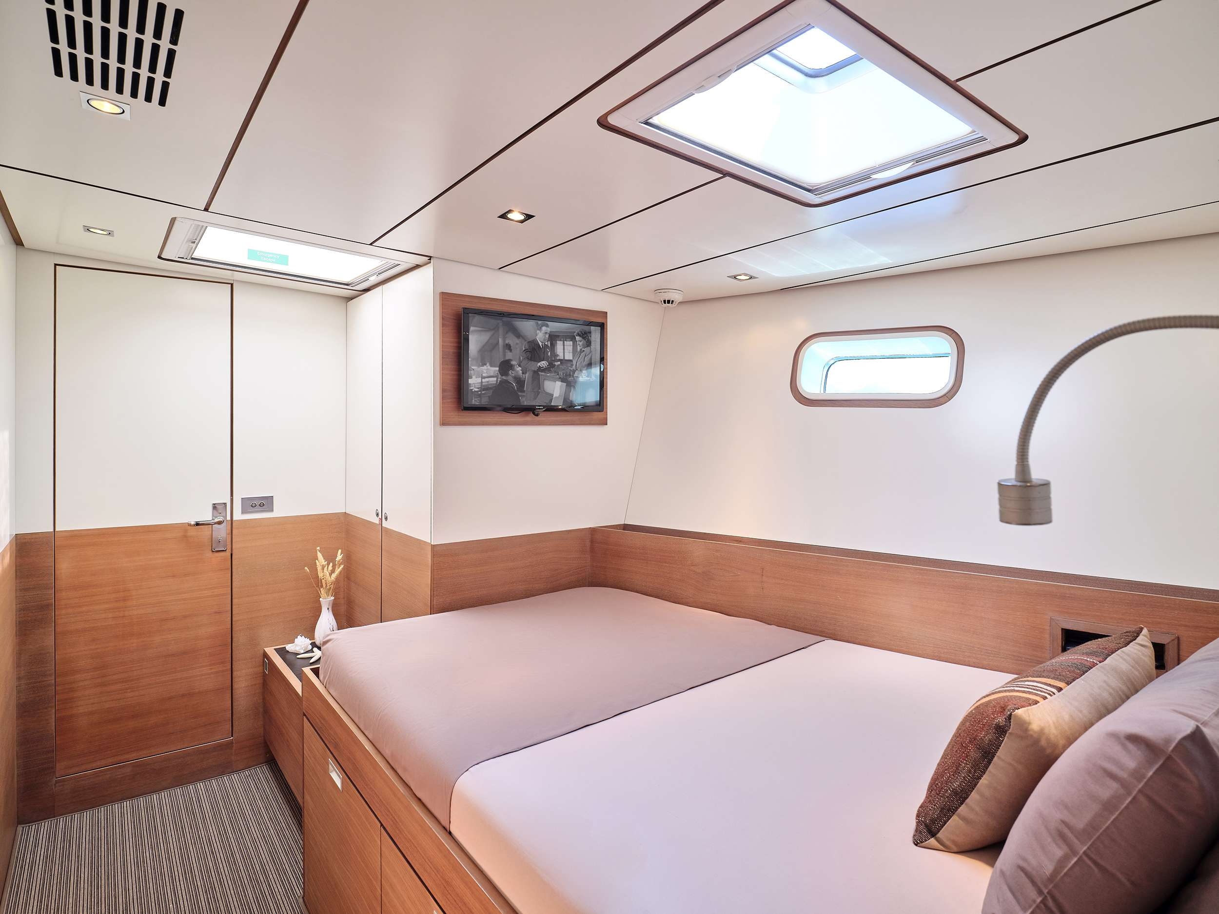 A modern yacht cabin available for yacht charter features wood paneling, a double bed, wall-mounted TV, small window, skylight, and minimal decor, including a vase with dried flowers on the bedside table.