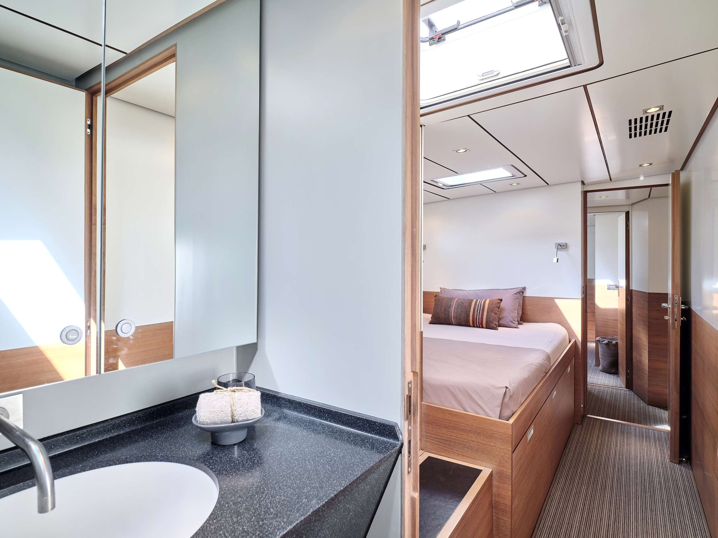 Modern yacht cabin interior with a compact bathroom featuring a sink and mirror, leading to a cozy bedroom with a double bed. Enjoy wood accents, natural light from a skylight, and contemporary fixtures—ideal for luxury yacht charter experiences.
