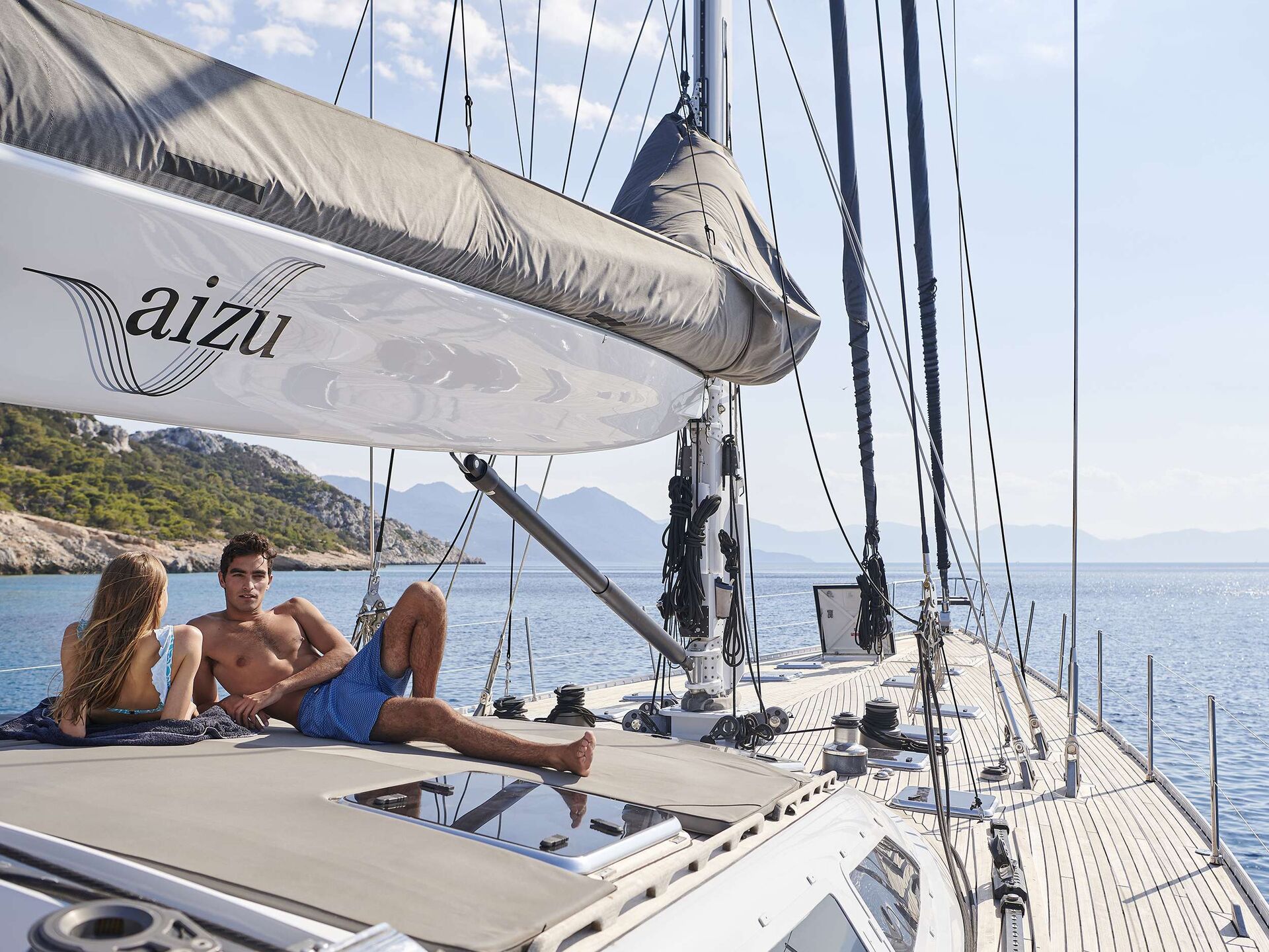 A man and woman relax on the deck of a sailboat named Aizu, anchored near a rocky coastline with clear blue water under a sunny sky—an ideal scene for those seeking premium yachts for charter.