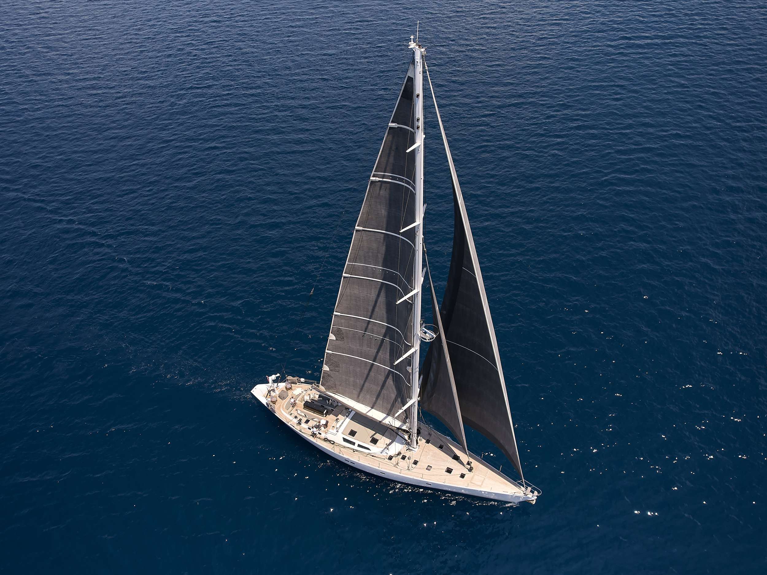 Aerial view of a sleek sailboat with dark sails gliding through calm, deep blue ocean waters—perfect for those seeking an unforgettable yacht rental experience.