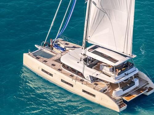 A large white catamaran yacht with sails up glides on bright blue ocean water. One person stands on the deck near the bow, enjoying the sunny, clear day at sea.