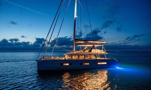 A modern catamaran yacht with interior and underwater lights glows on calm water at dusk, with a dramatic, cloud-filled sky in the background.