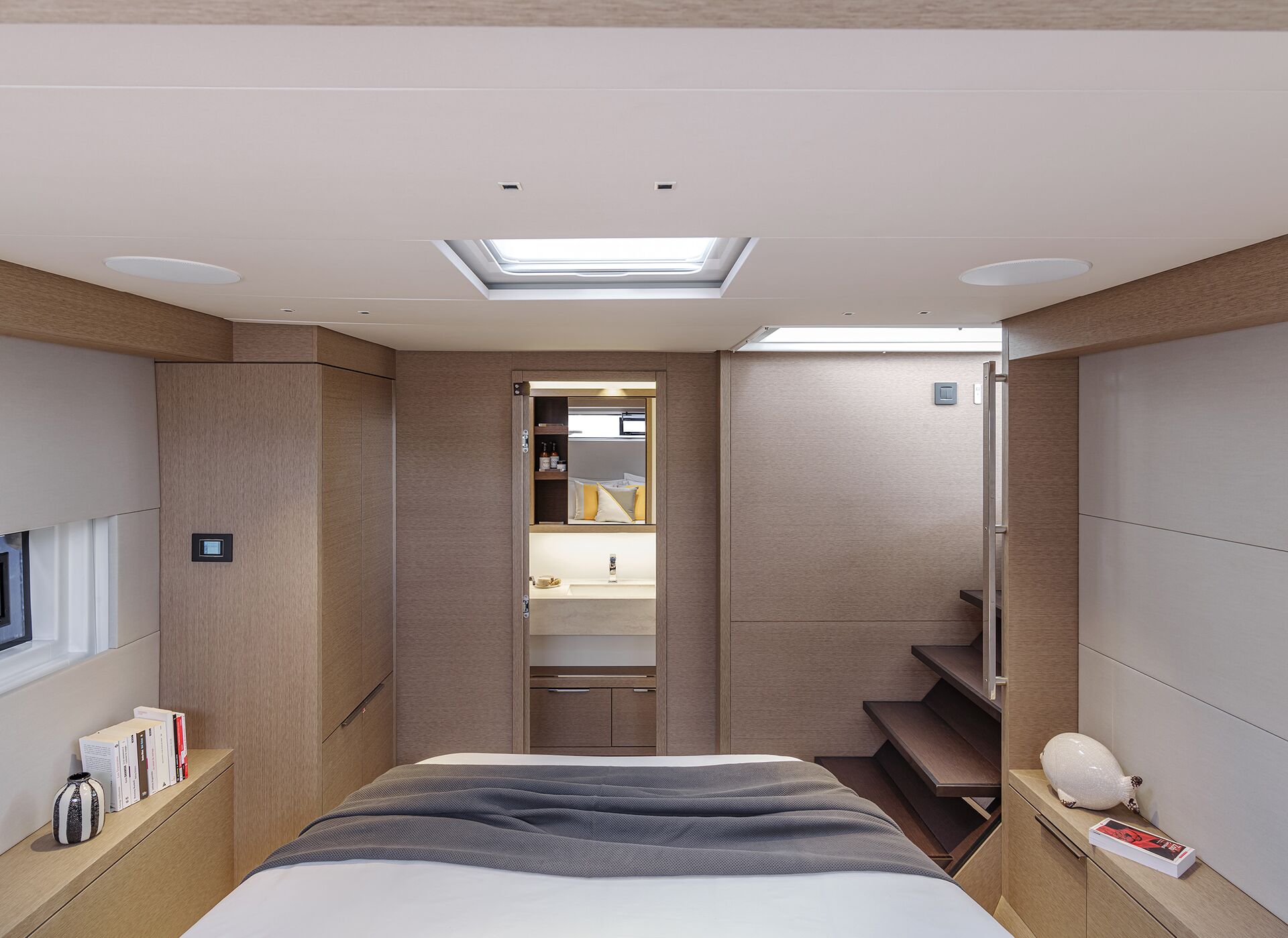 A modern yacht bedroom with a double bed, light wood paneling, a skylight, shelves with books, a small sink area beyond the foot of the bed, and stairs leading up to the right.