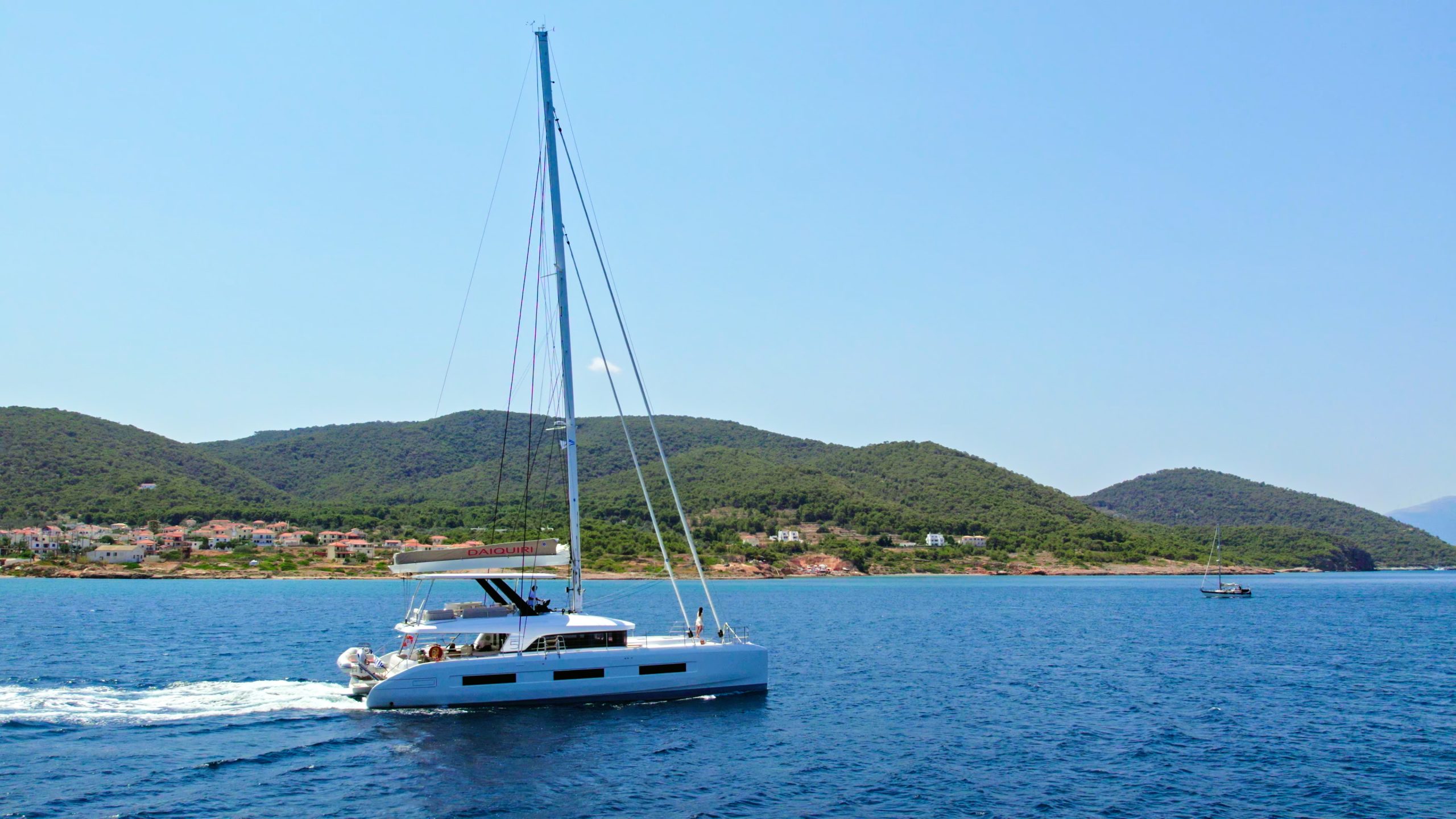 A white catamaran yacht sails on calm blue water near a green, hilly coastline under a clear sky, with a small town and another boat visible in the distance.