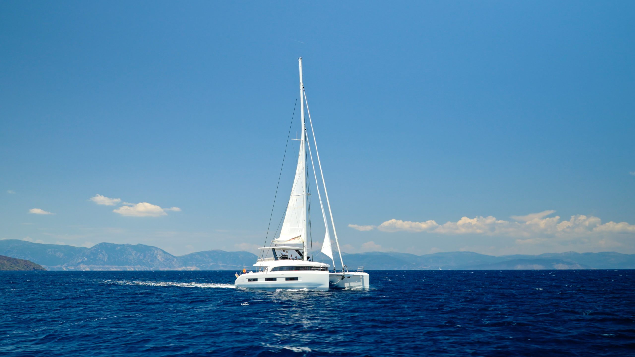 A white catamaran yacht with raised sails cruises on deep blue water under a clear sky, with distant mountains and a few clouds visible on the horizon.