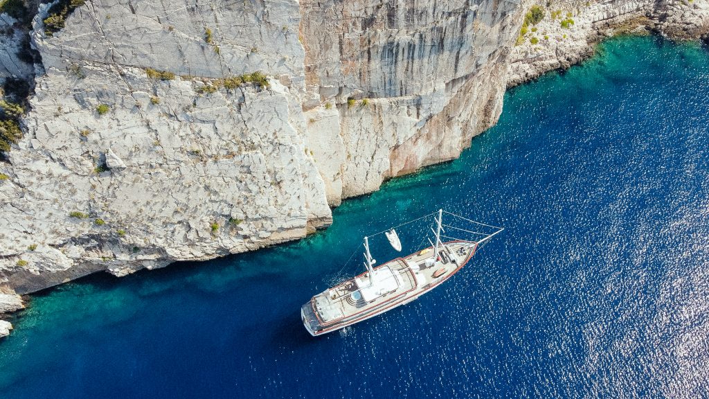 Aerial view of a sailboat anchored in clear blue water near a tall rocky cliff with sparse vegetation. The sunlight highlights the textures of the rock and the vibrant color of the sea.