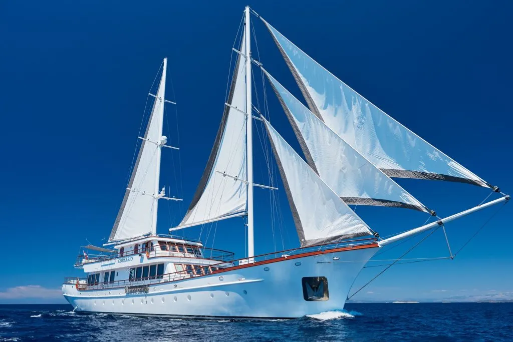 A large white sailing yacht with two masts and several sails open, cruising on calm blue water under a clear sky.