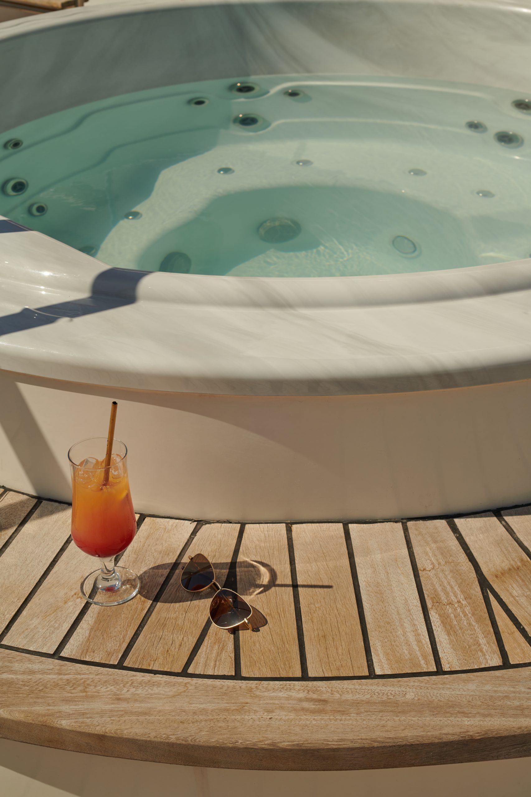 A tropical drink with a straw and a pair of sunglasses rest on wooden decking beside a bubbling hot tub filled with clear water.